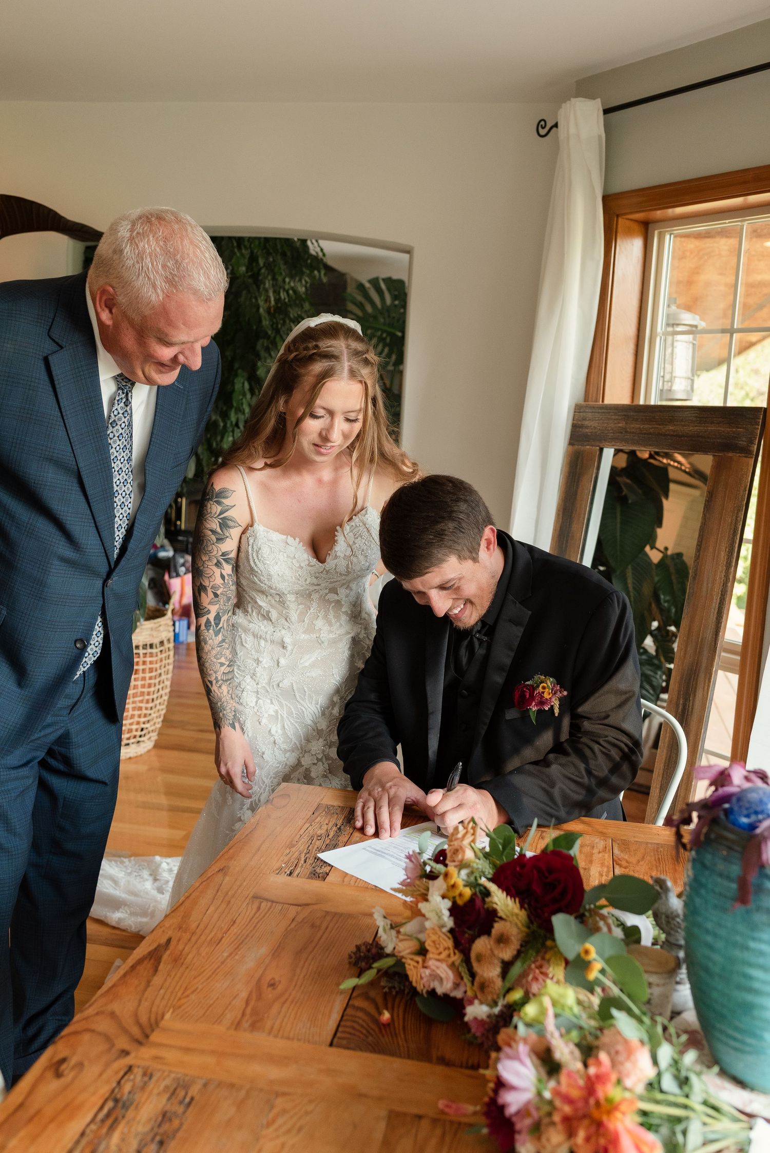 Bride and groom signing wedding marriage certificate at wooden table with floral bouquet, witnessed by officiant indoors.