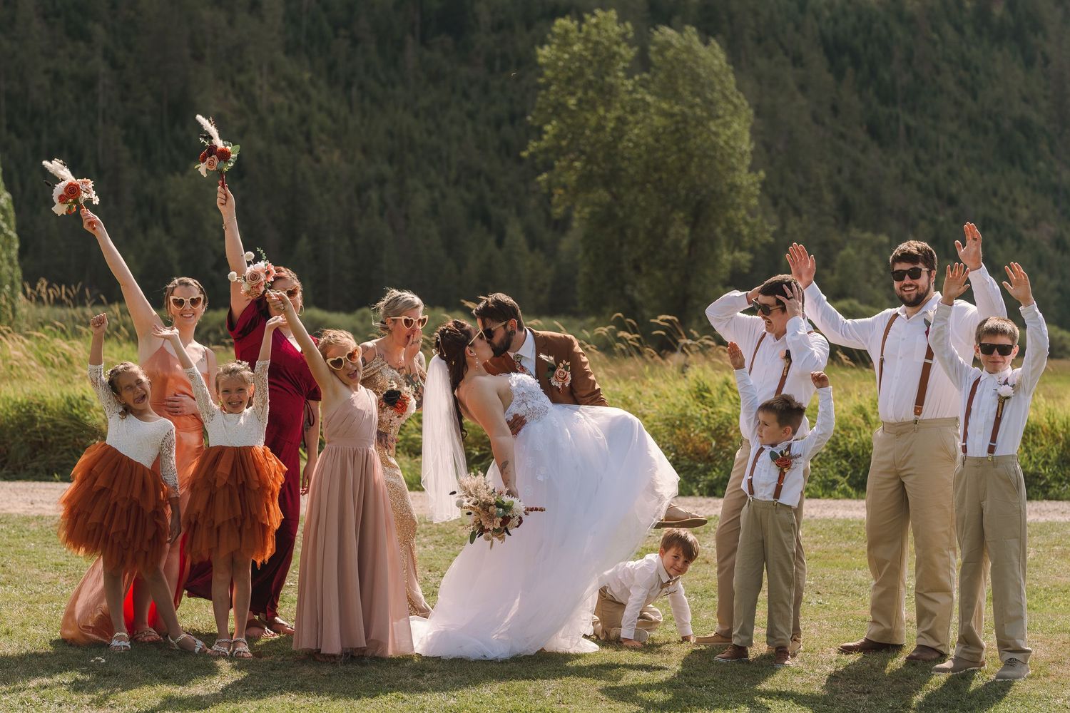 Wedding party celebrating outdoors, bridesmaids in pink and rust dresses, groomsmen in tan suits, bride and groom kissing.