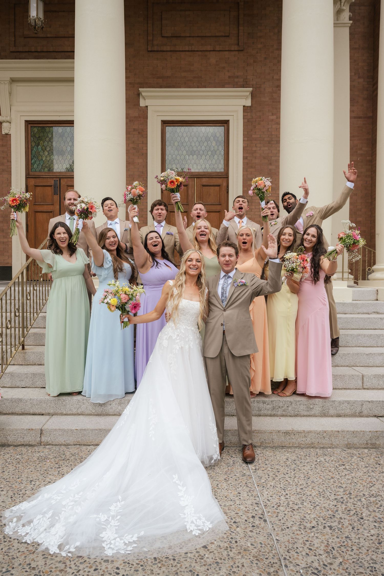 Bride and groom pose with cheering wedding party in colorful gowns on brick building steps.