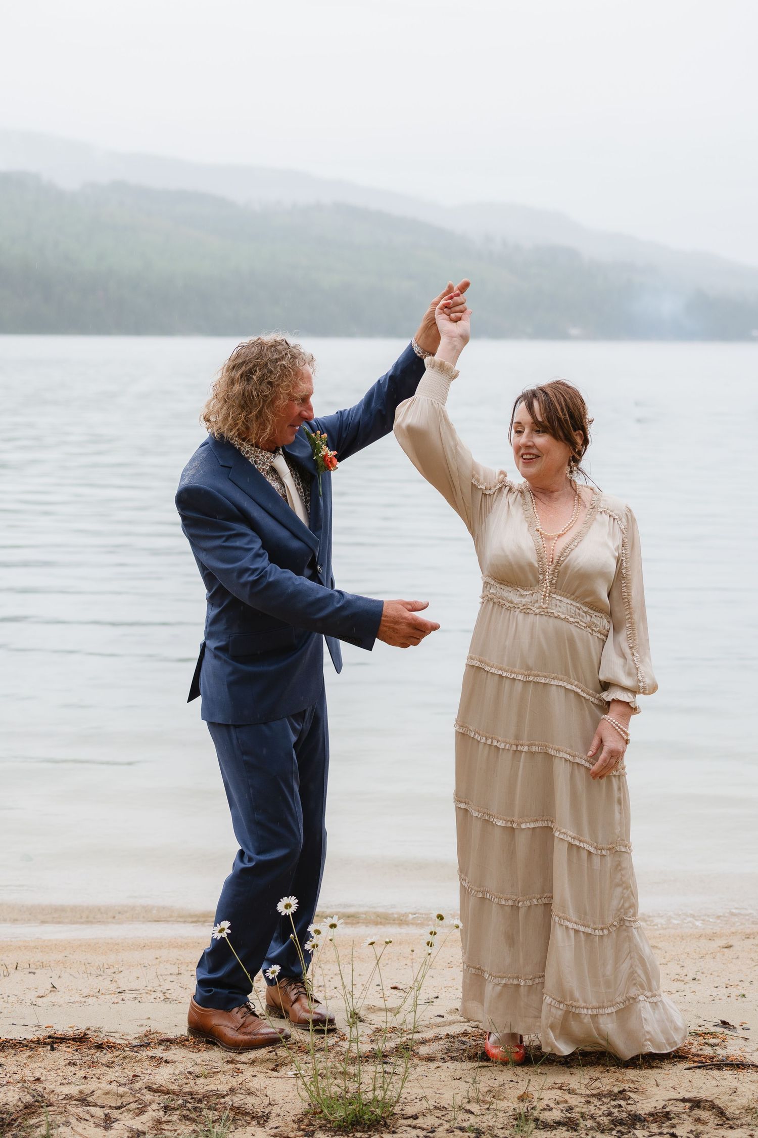 A couple dances joyfully together on a misty beach with mountains visible in the background.