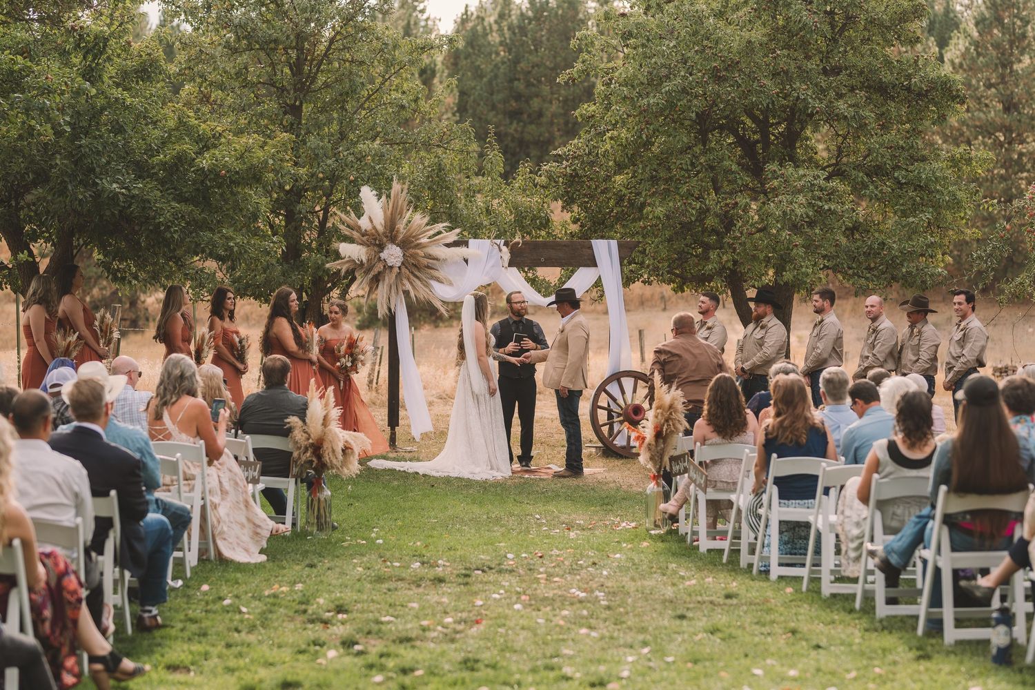 Panoramic view of an outdoor wedding ceremony with guests seated in white chairs on a grassy lawn surrounded by trees.
