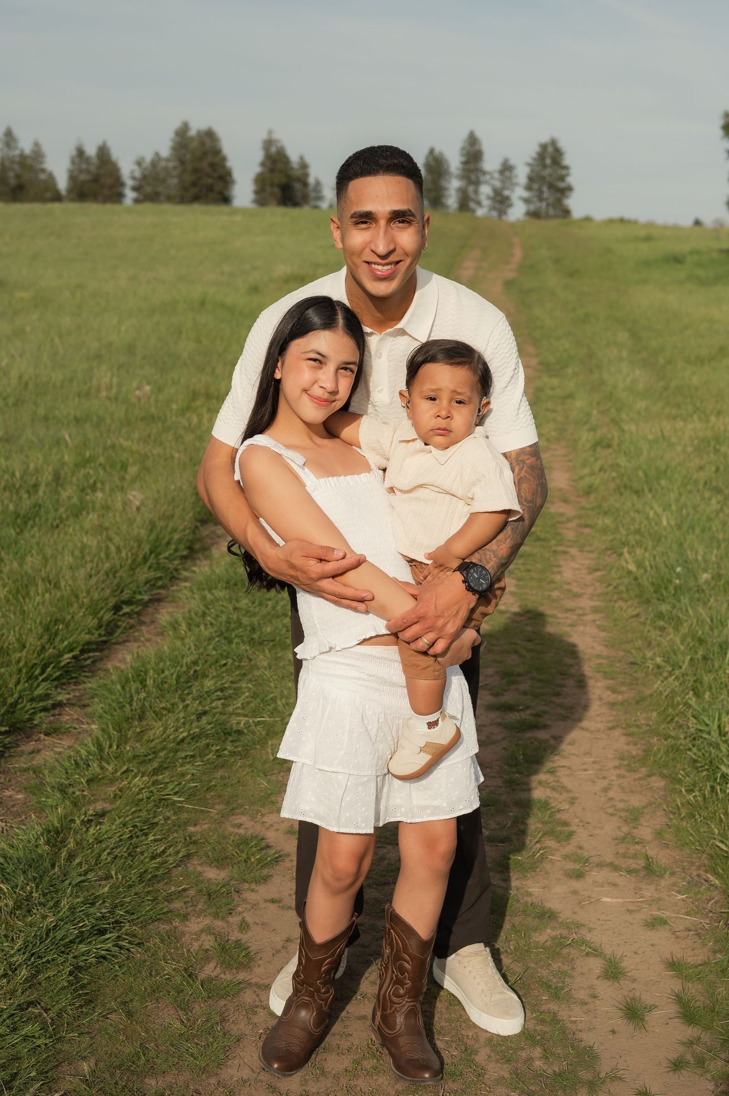 Young family of three posing on a grassy field path, parents in white outfits holding their baby, with trees in background.