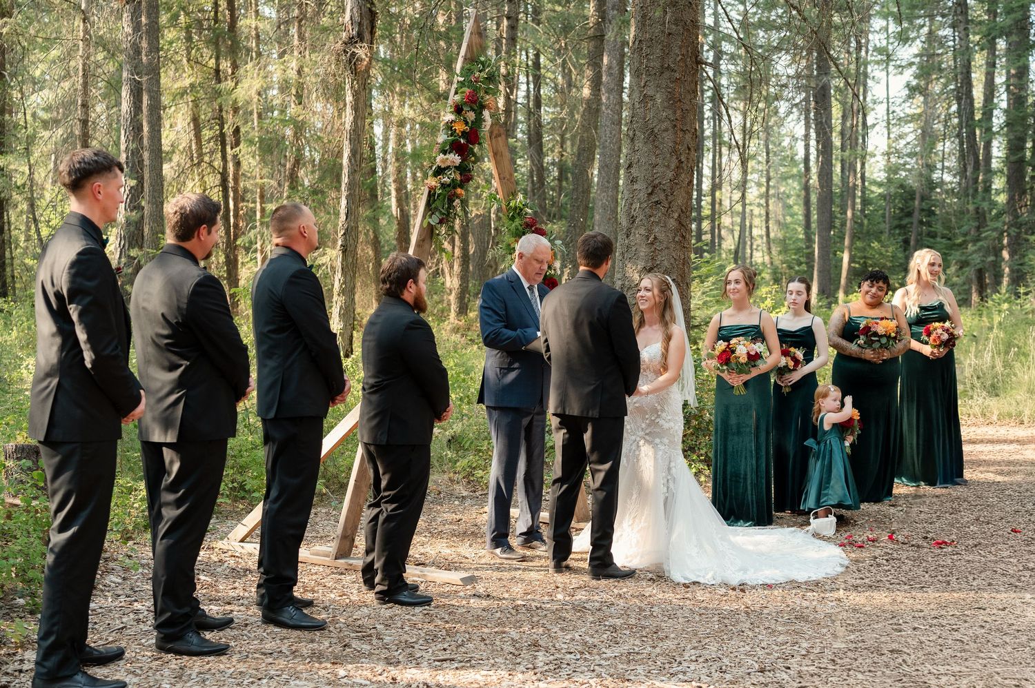 Outdoor wedding ceremony in a forest with bride, groom, officiant, groomsmen in black suits, and bridesmaids in green dresses.