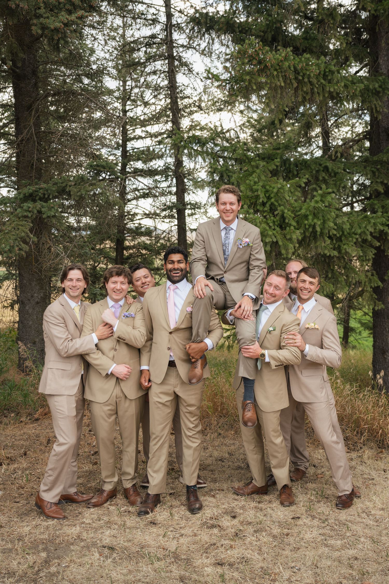 Groomsmen in tan suits pose outdoors near pine trees, lifting the groom in a fun wedding party photo.