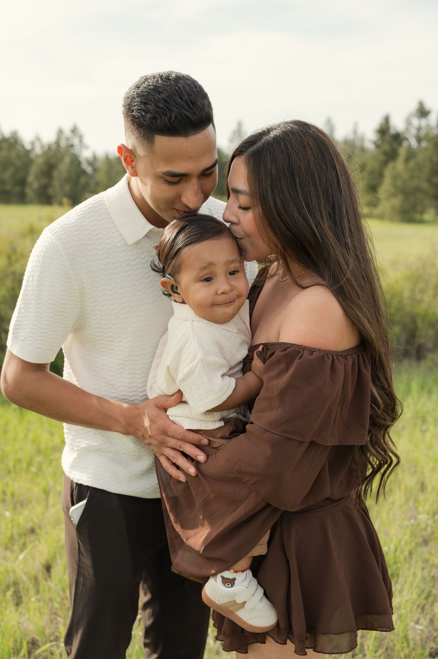 A young family poses outdoors in a green field, parents kissing their smiling baby dressed in white.