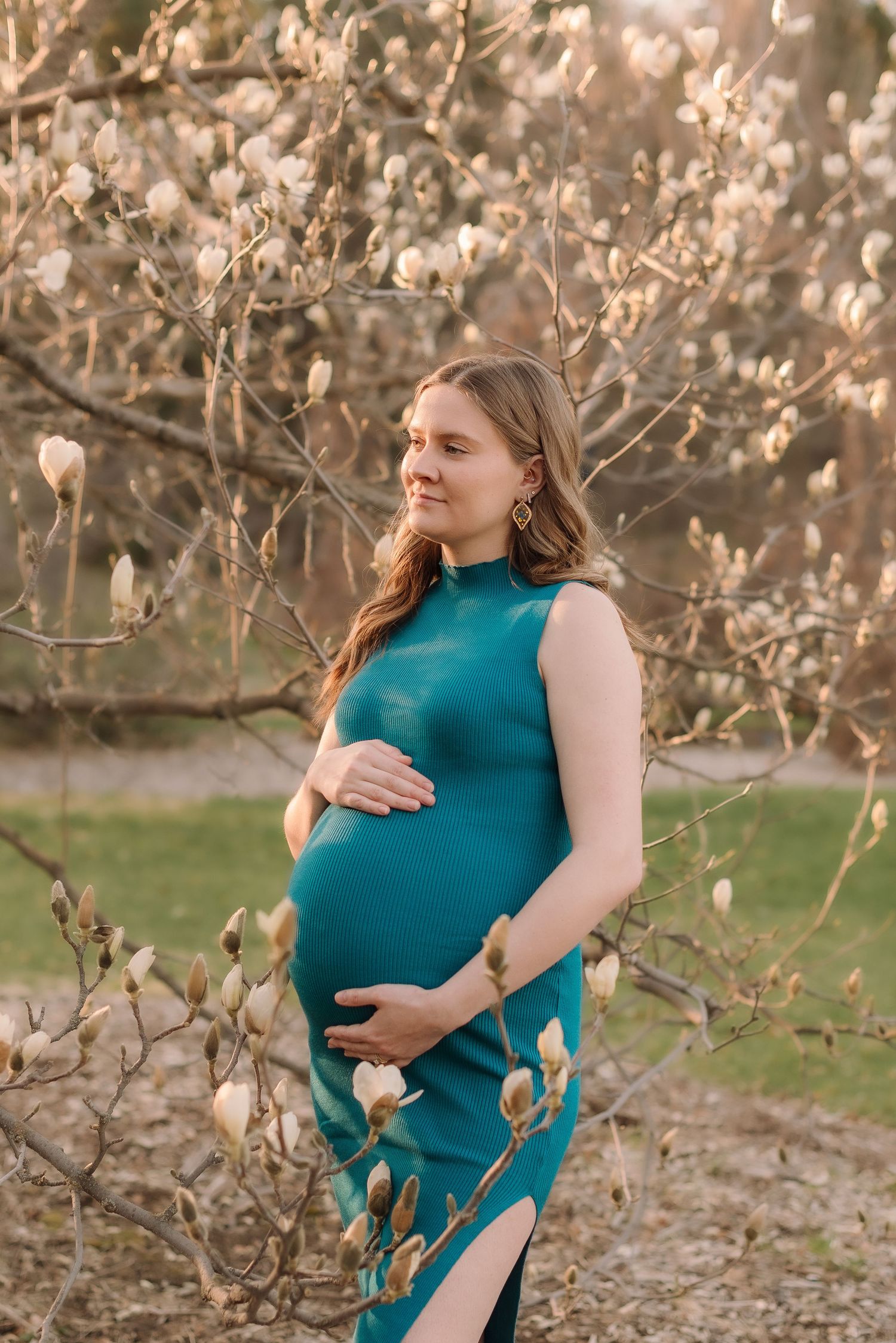A pregnant woman in a teal dress stands among blooming spring tree branches at sunset.