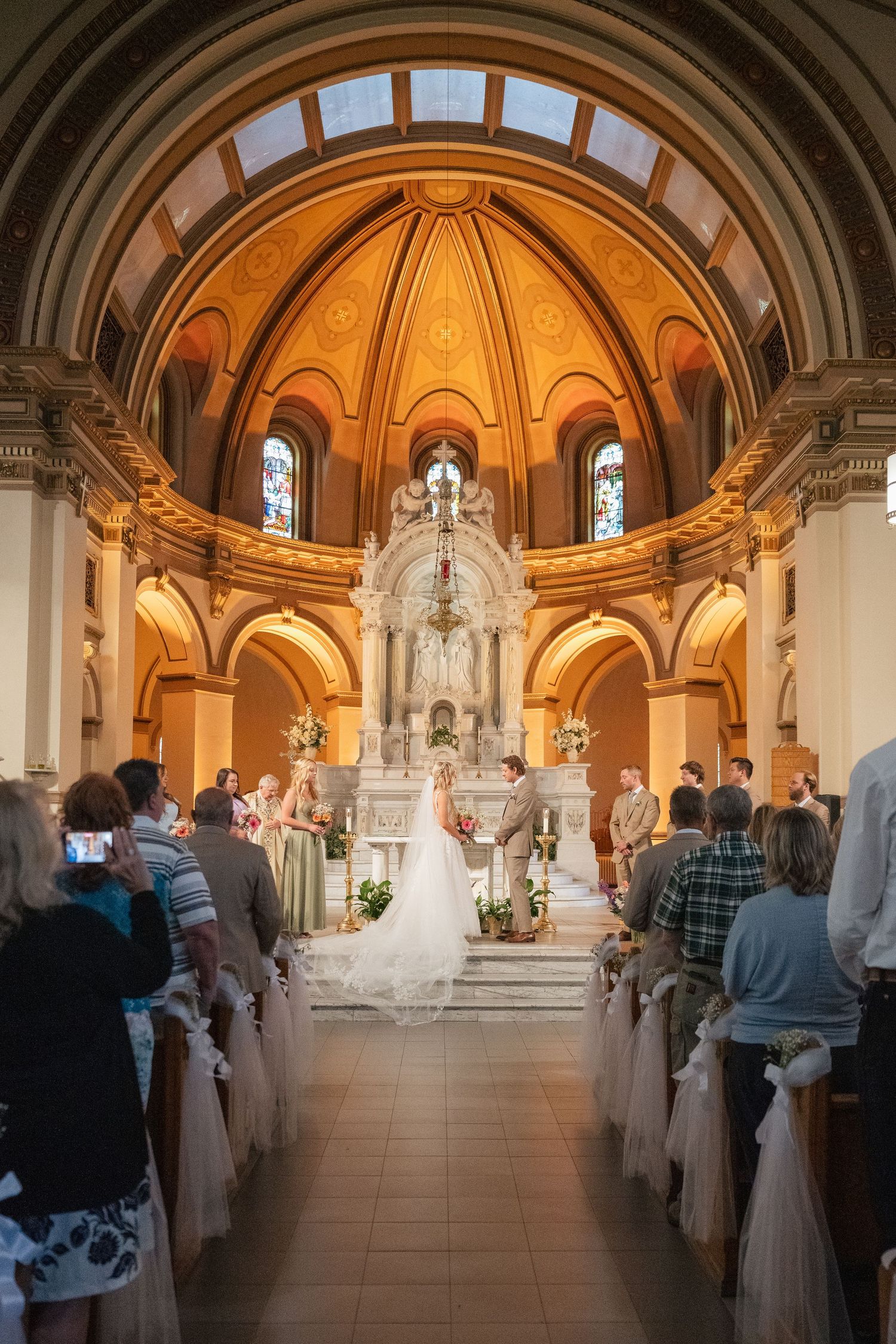 Elegant wedding ceremony in a grand church with ornate golden arches, bride and groom at altar, guests seated in decorated pews.