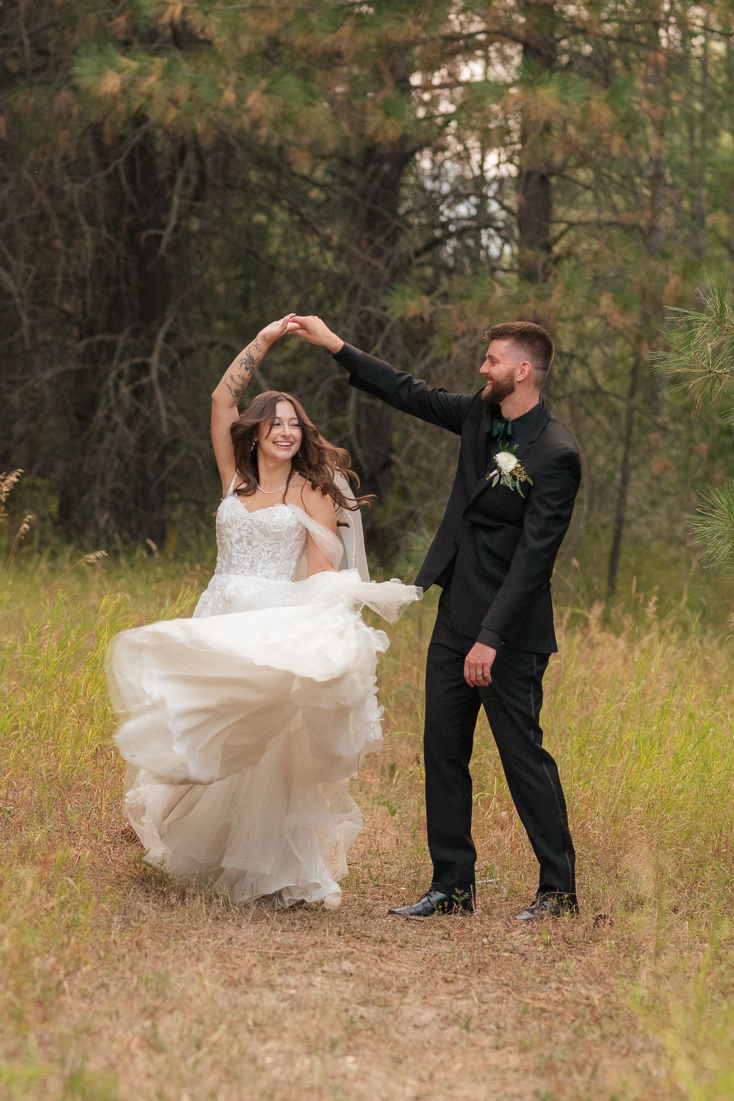 Bride in white gown twirls as groom in black suit holds her hand, dancing outdoors in a forest meadow.