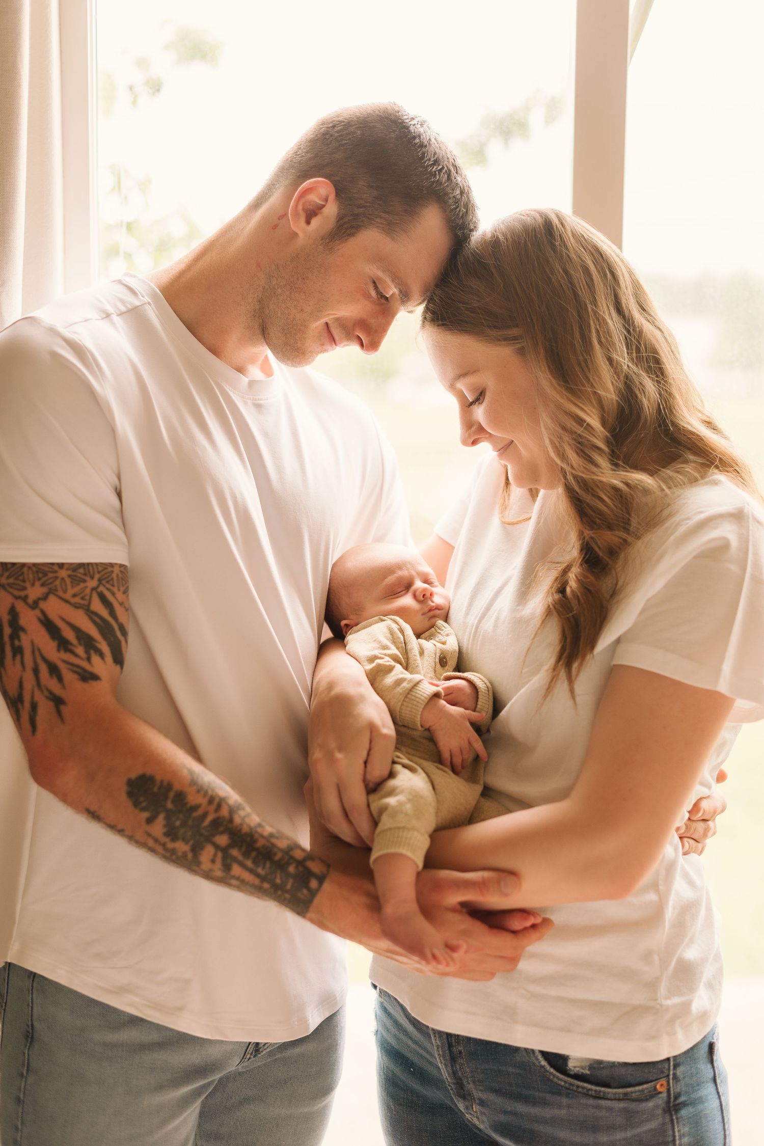 A couple in white t-shirts and jeans share tender moments with their newborn baby in a bright sunlit room.