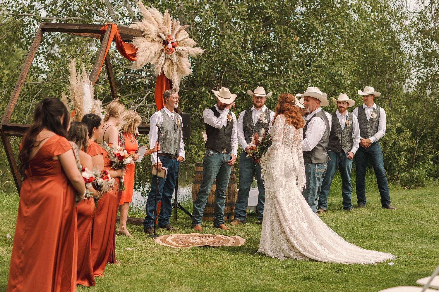 A rustic outdoor wedding ceremony with bridesmaids in coral dresses and groomsmen in cowboy hats under a floral arch.