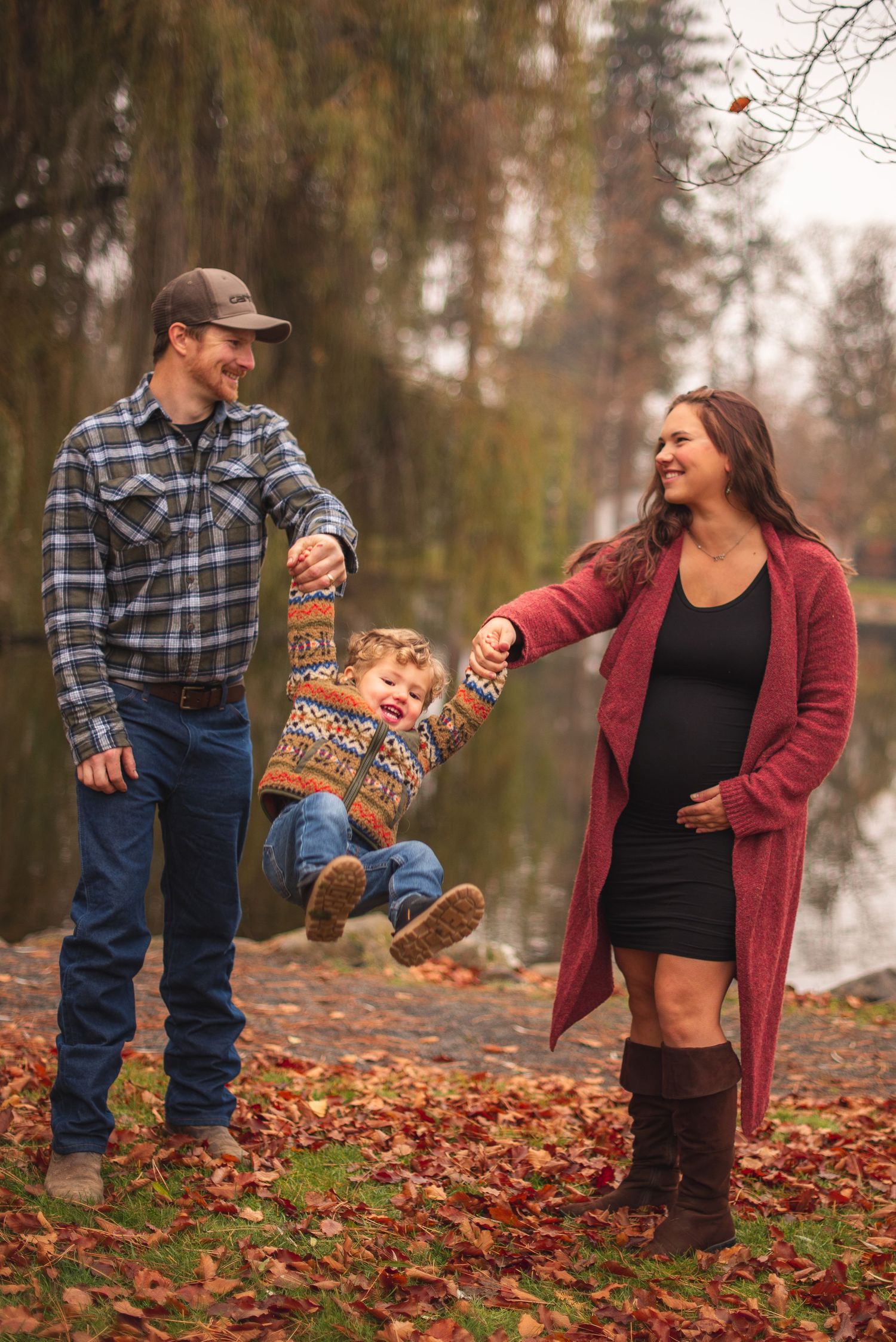 Parents swinging toddler between them on path covered in fall leaves.