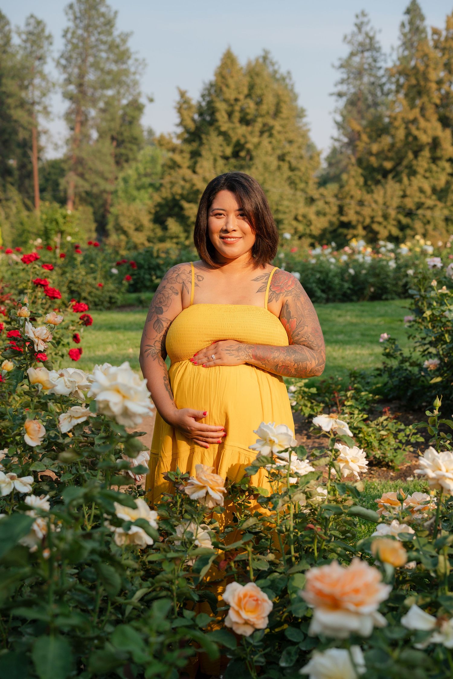 Pregnant woman in yellow dress cradling baby bump among blooming roses in a lush garden park.