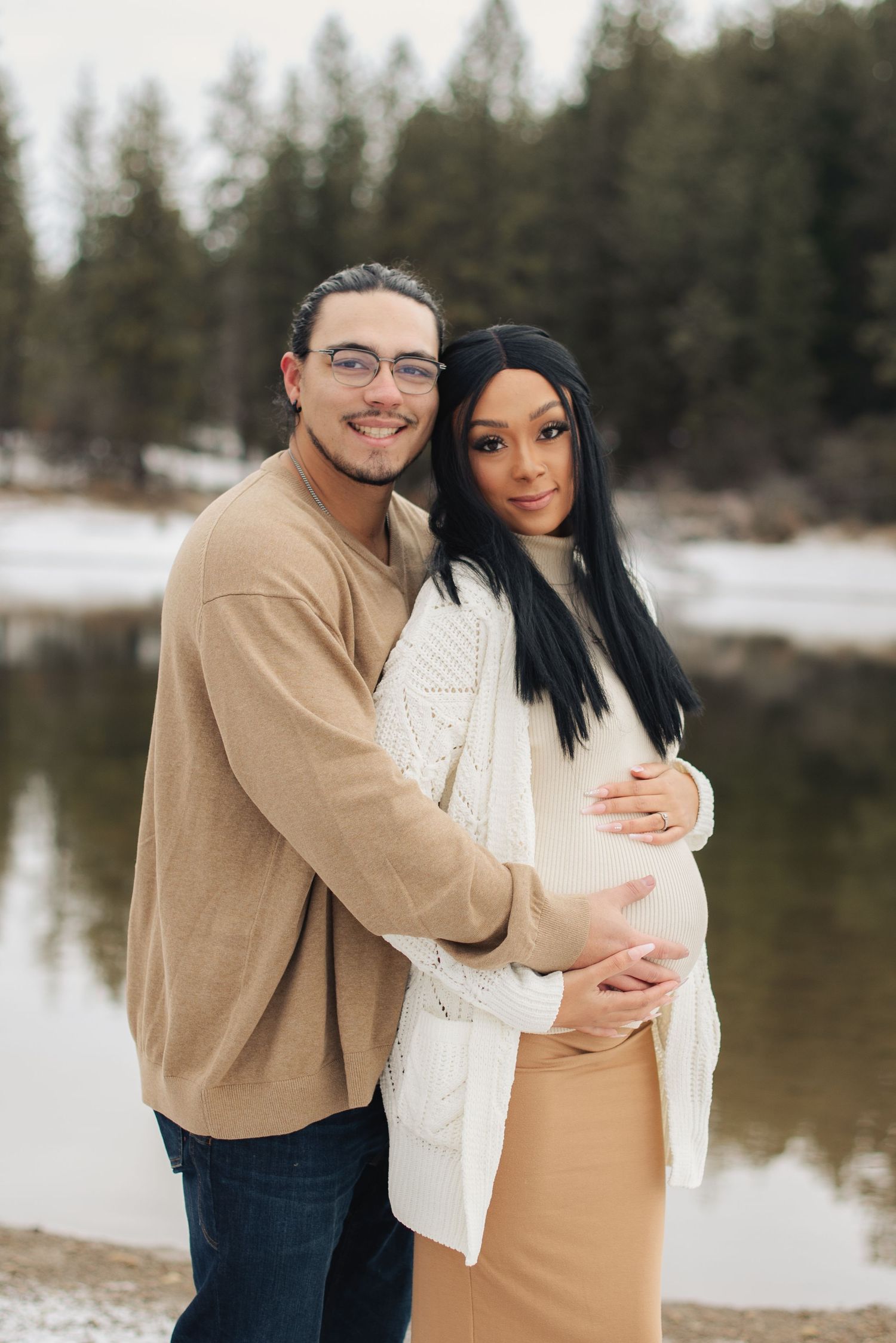 A couple poses outdoors in winter, the woman visibly pregnant, both smiling warmly near a snowy lake with pine trees.