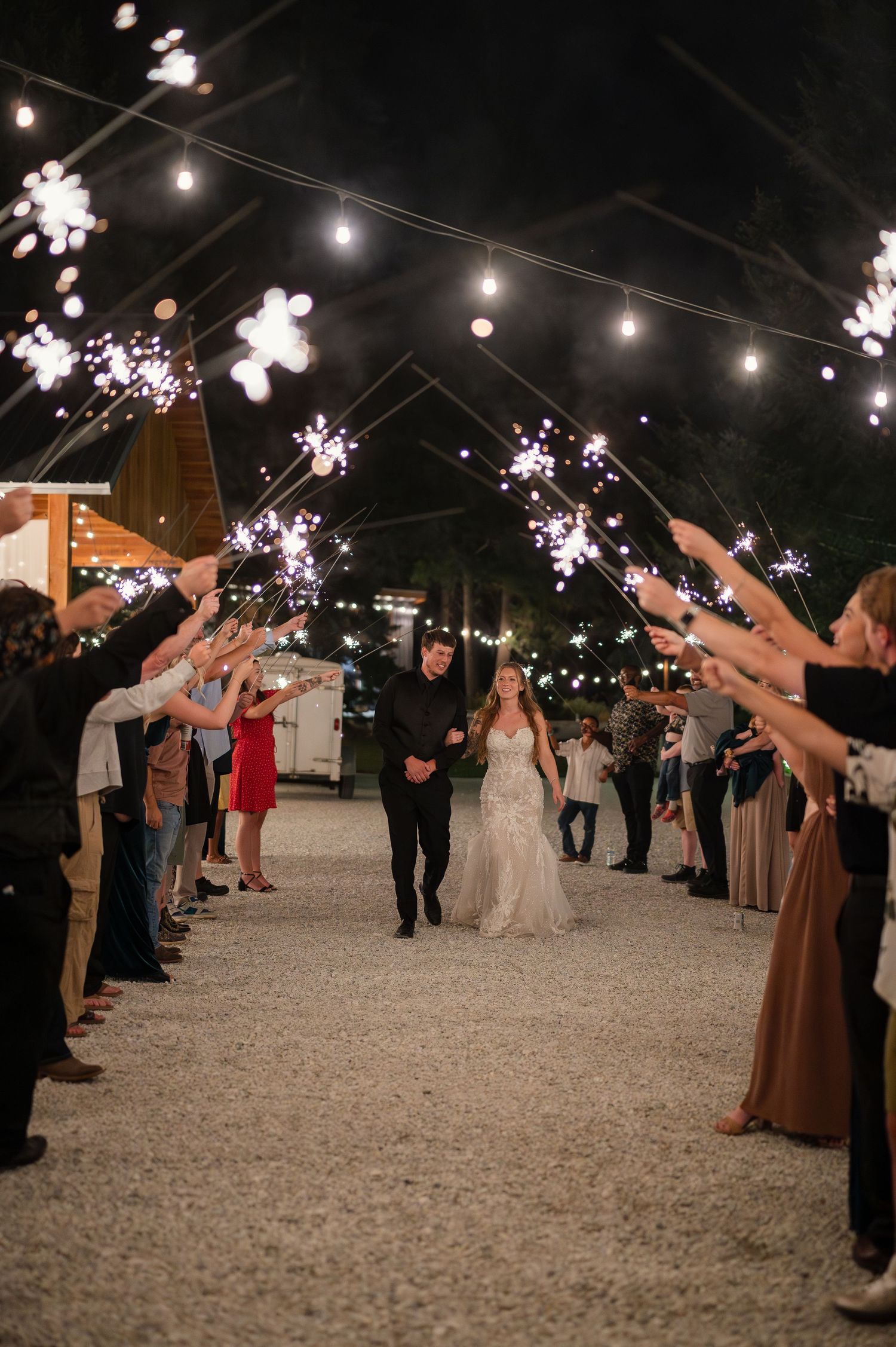 Bride and groom walk through sparkler tunnel at night wedding reception with string lights overhead.