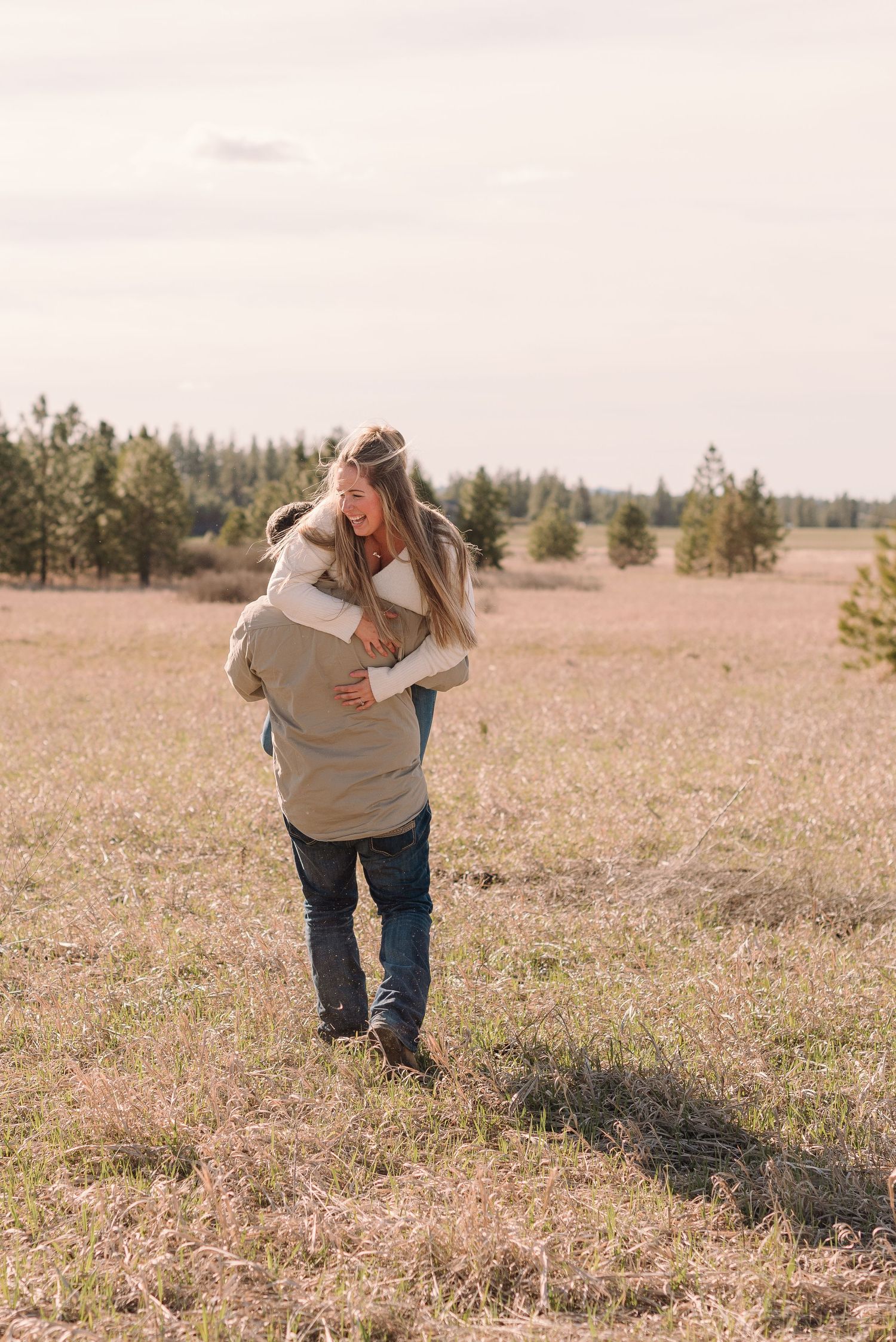 Two people share a tender embrace while standing in a golden field with evergreen trees in the background.