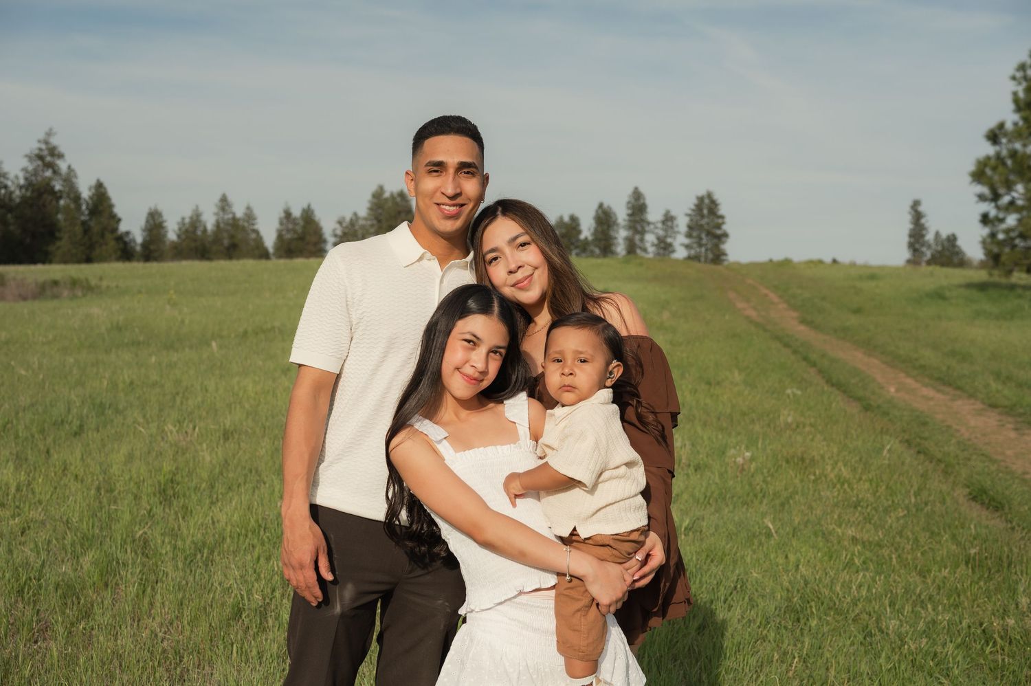 Family of four posing in a green meadow, parents holding children, smiling outdoors on a sunny day.