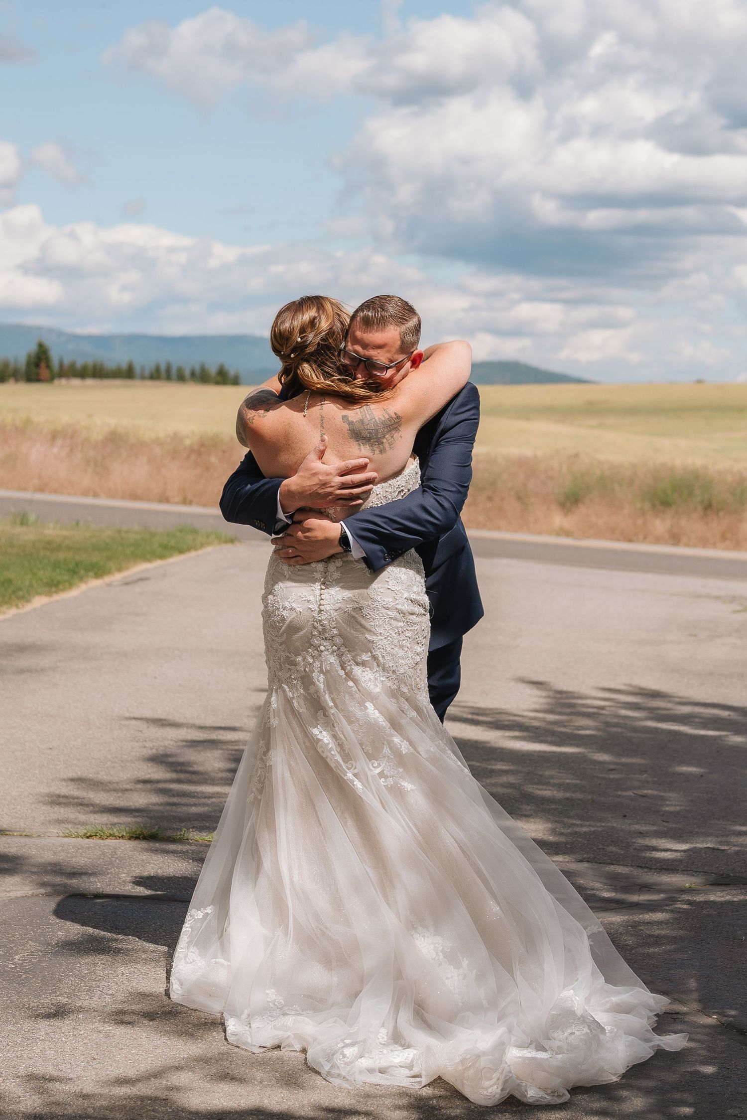 A couple in wedding attire share an intimate dance on a rural road with mountains and clouds in the background.