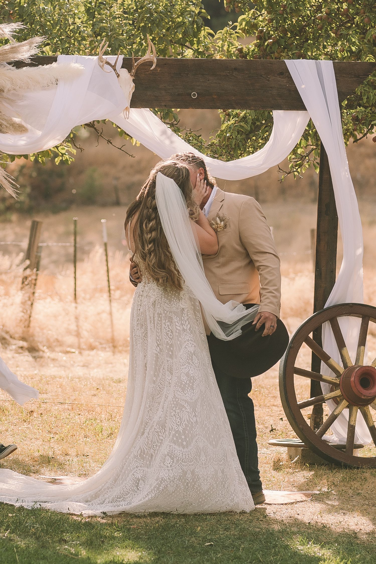 Romantic wedding couple embraces near rustic wagon wheel with flowing white veil in outdoor garden setting.