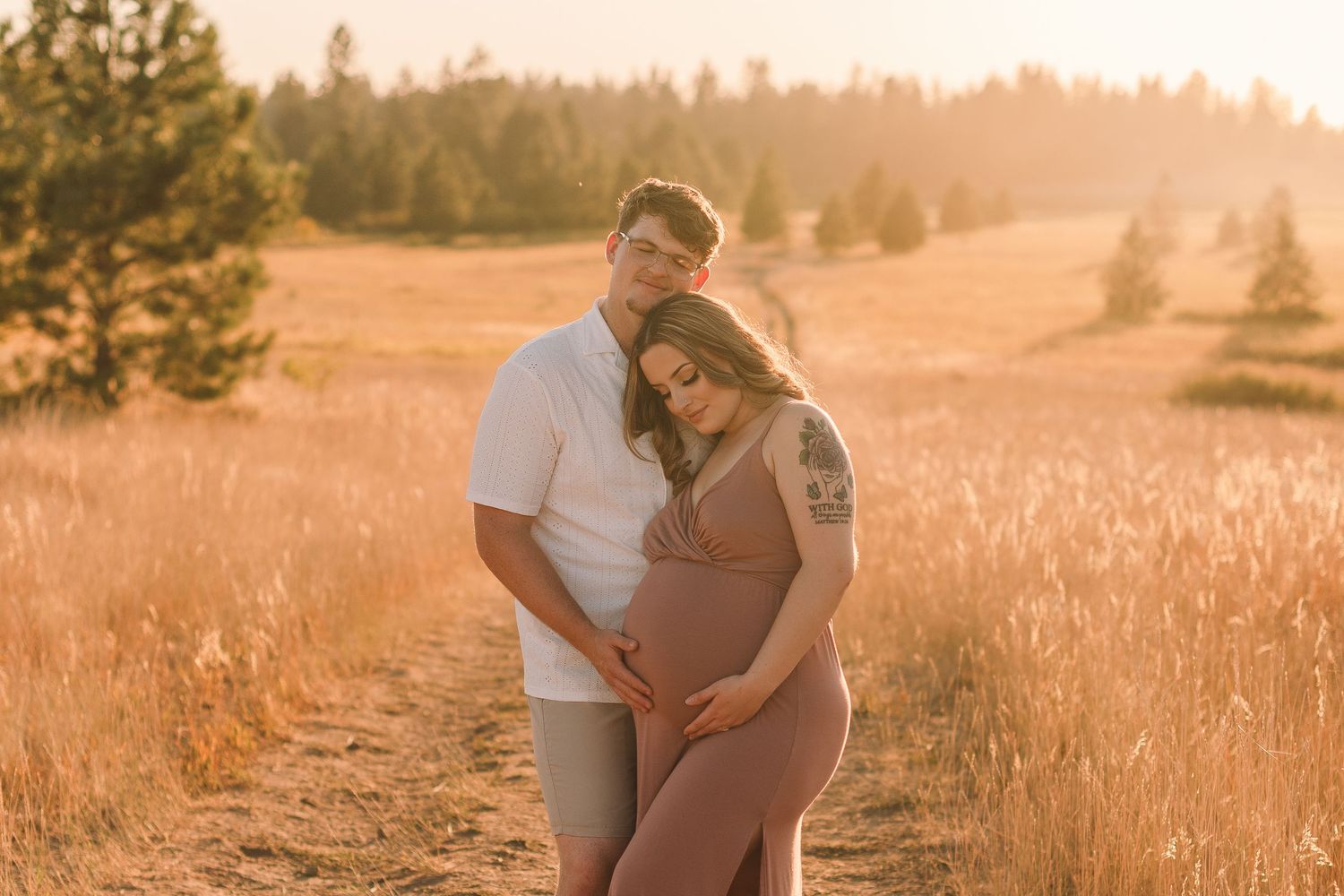 Couple embraces in a golden wheat field at sunset with tall pine trees in the background.