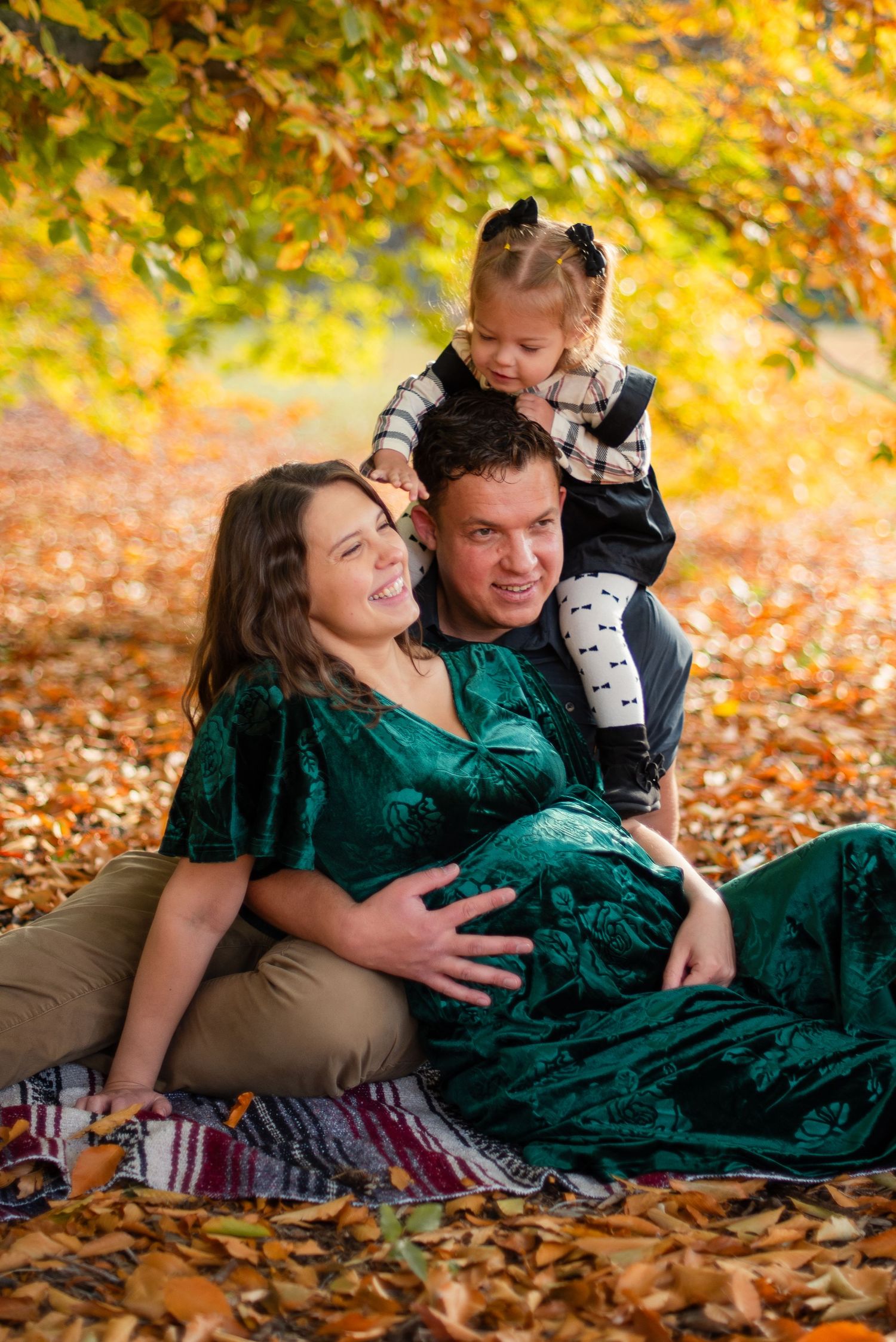Pregnant woman in green dress with husband and toddler daughter enjoying autumn family maternity photo session outdoors.