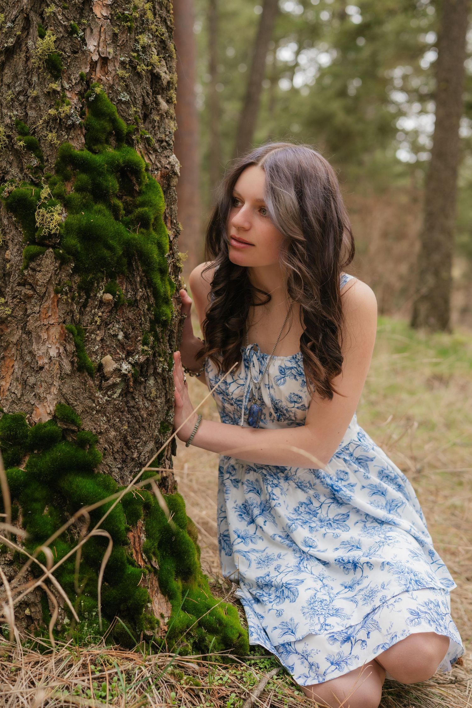 A person in a blue and white floral dress kneels beside a mossy tree trunk in a dreamy forest setting.