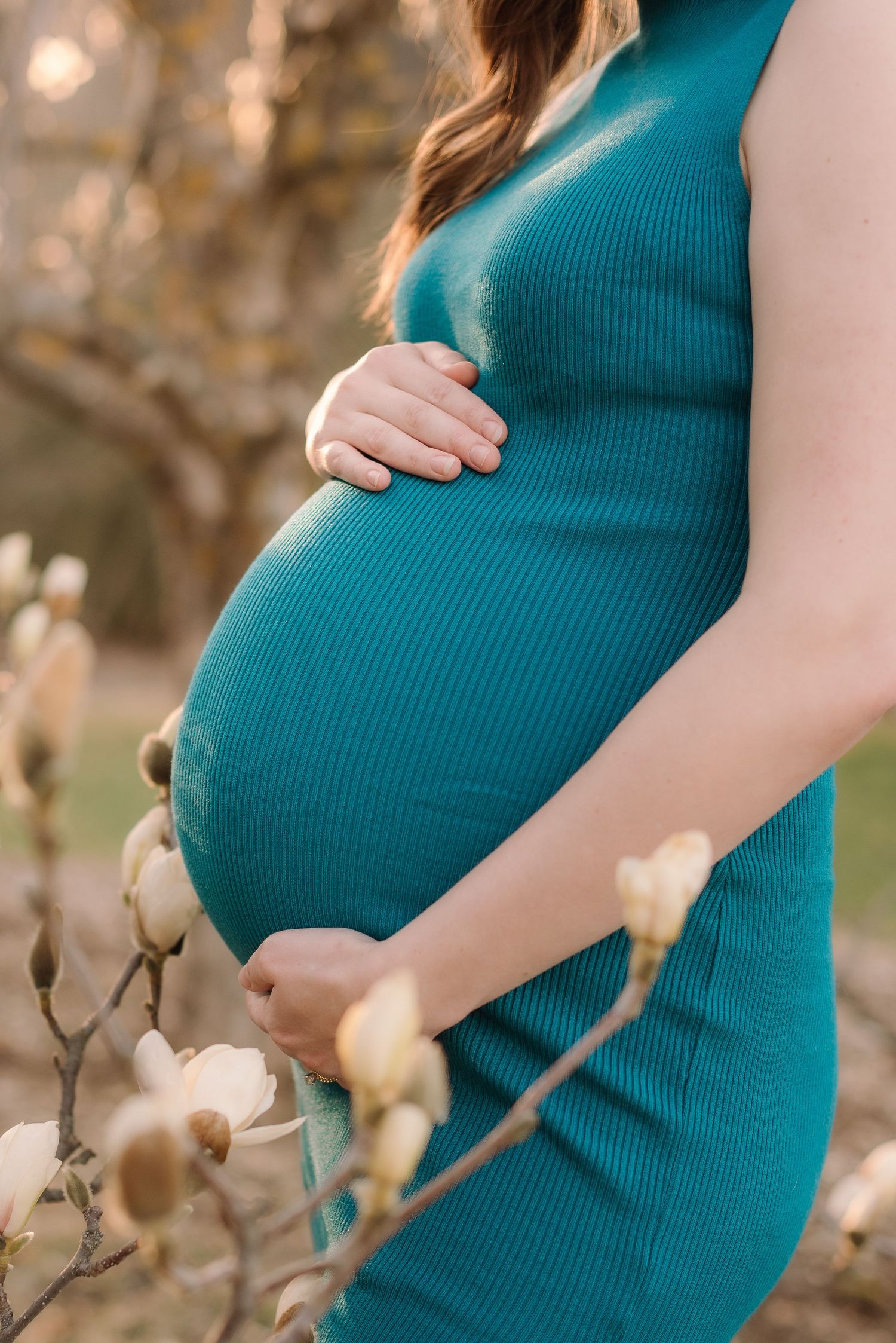 A pregnant woman in a teal ribbed dress cradles her baby bump while standing among flowering magnolia branches.