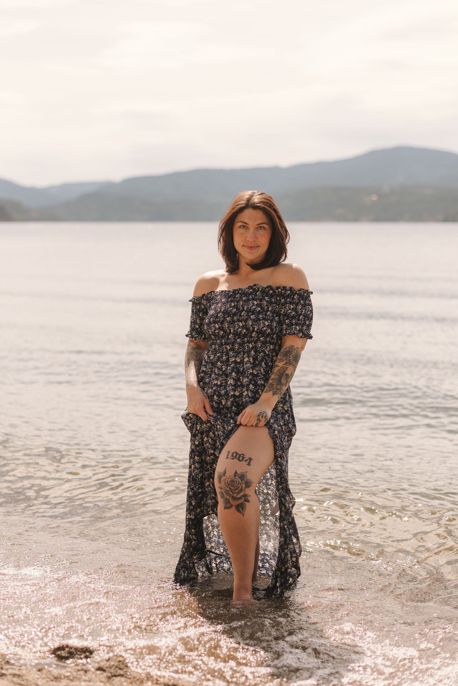 Woman in floral dress poses gracefully in shallow ocean water with mountains visible in background.