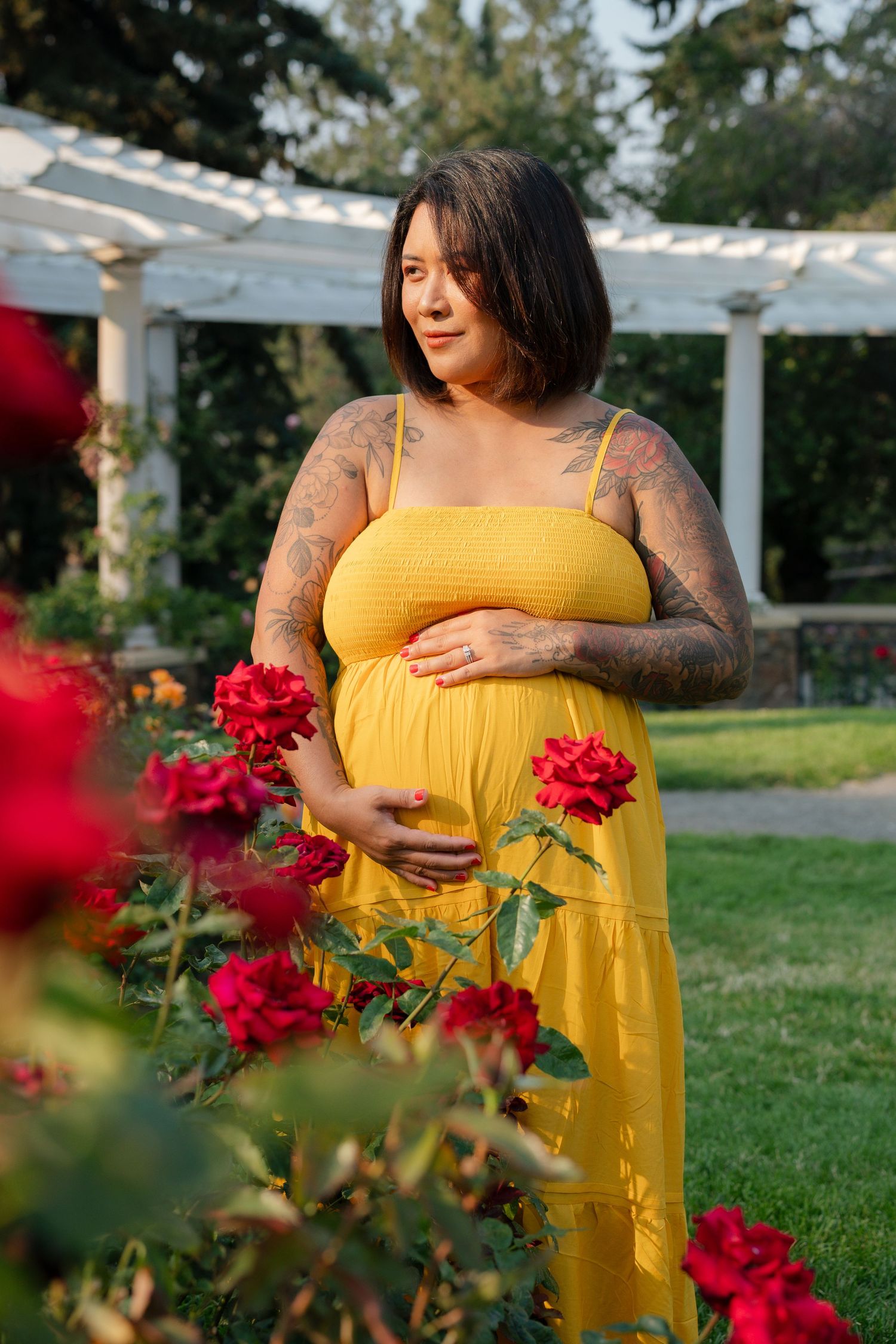 Pregnant woman in yellow dress with tattoos cradling baby bump among red roses in a garden.