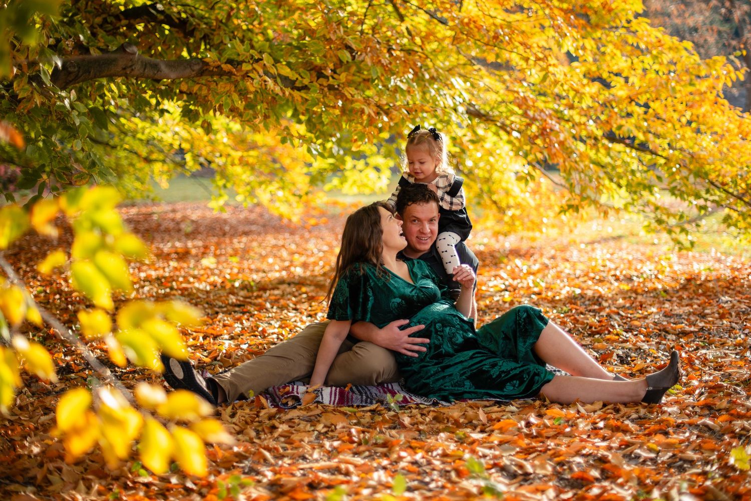 Family sitting on autumn leaves under golden trees, pregnant mother in green dress with partner and toddler.