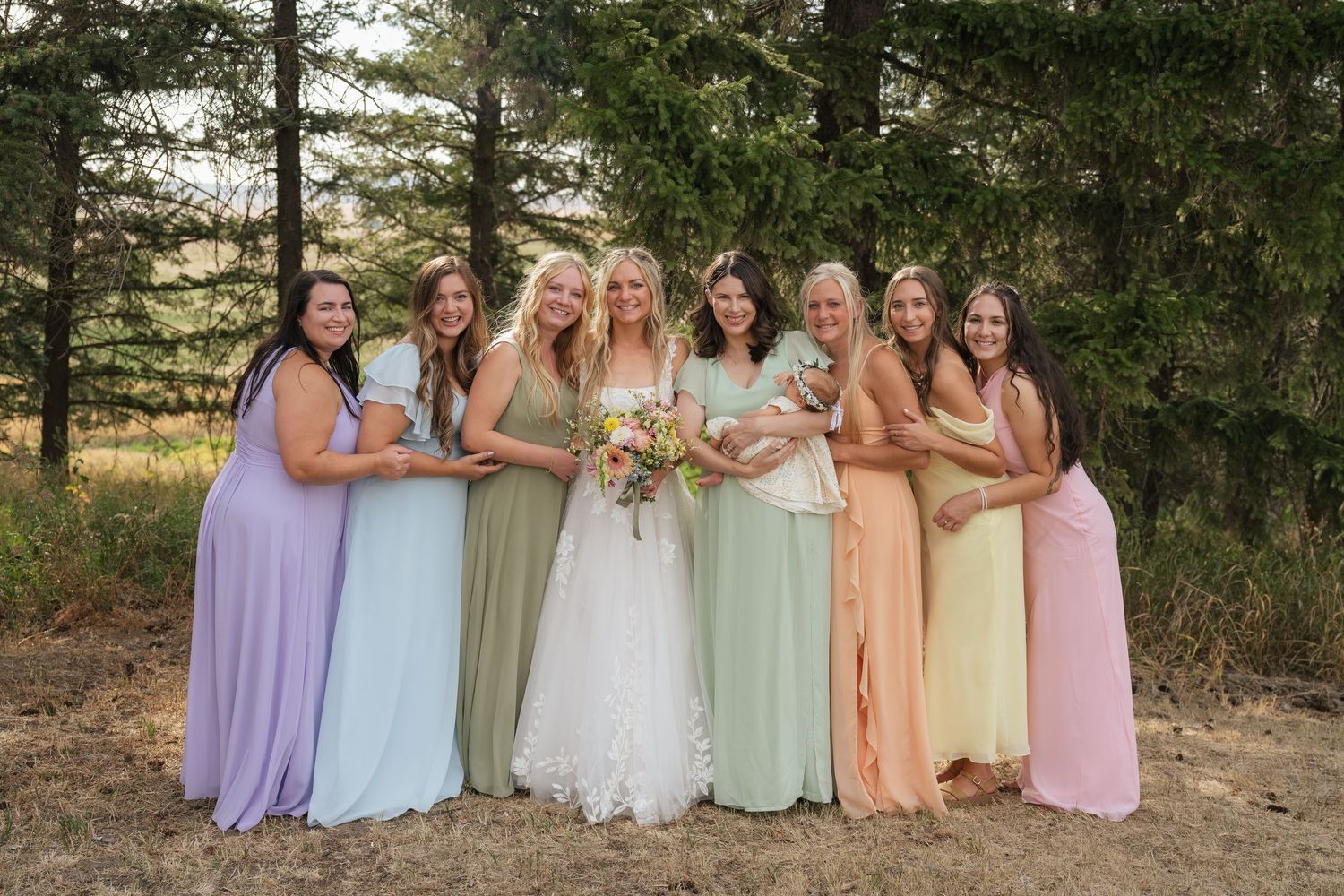 Bride in white gown poses with seven bridesmaids in colorful mismatched dresses outdoors among pine trees.