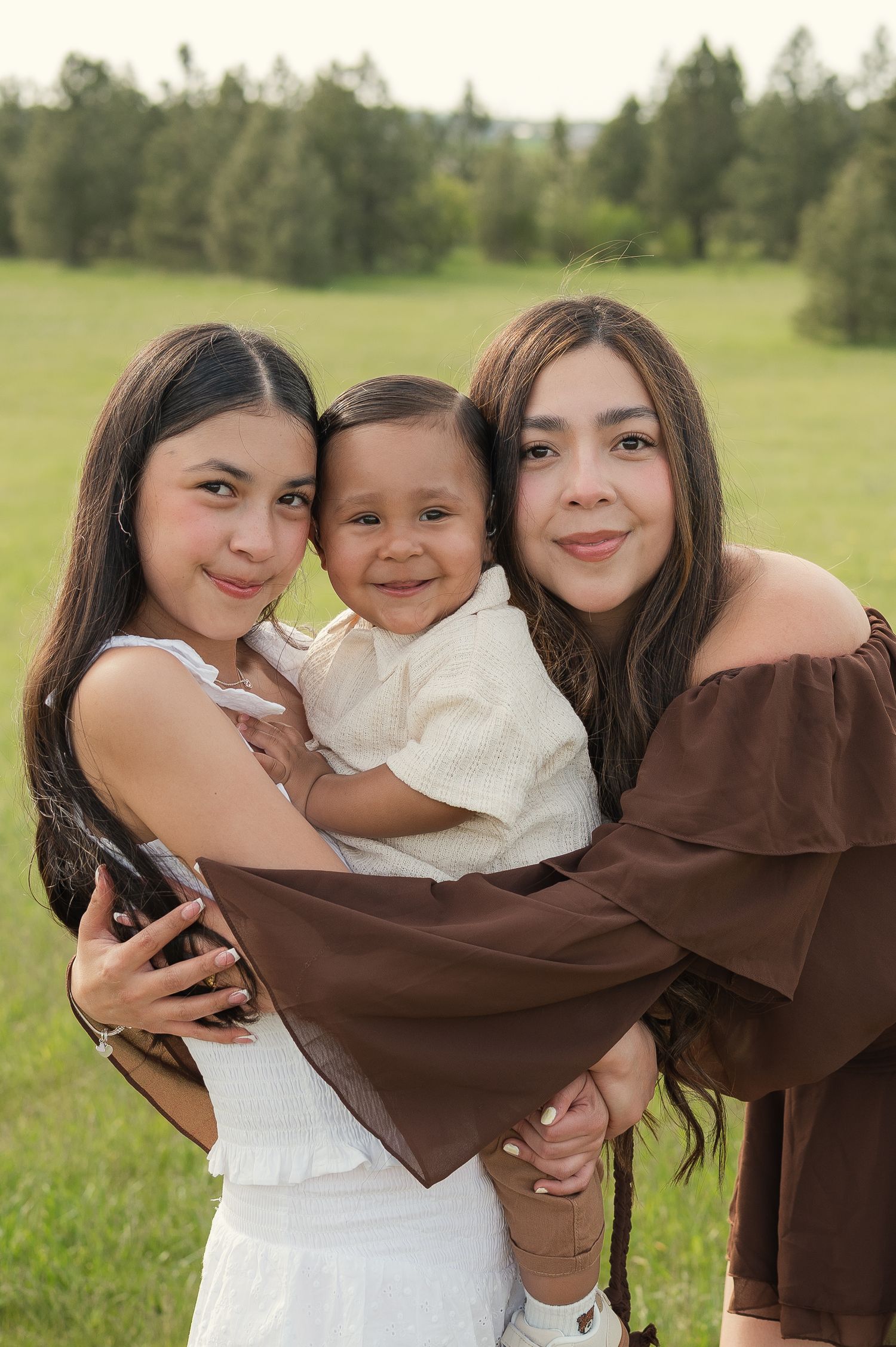 Two women and a smiling baby boy pose together in a green field, dressed in white and brown outfits.