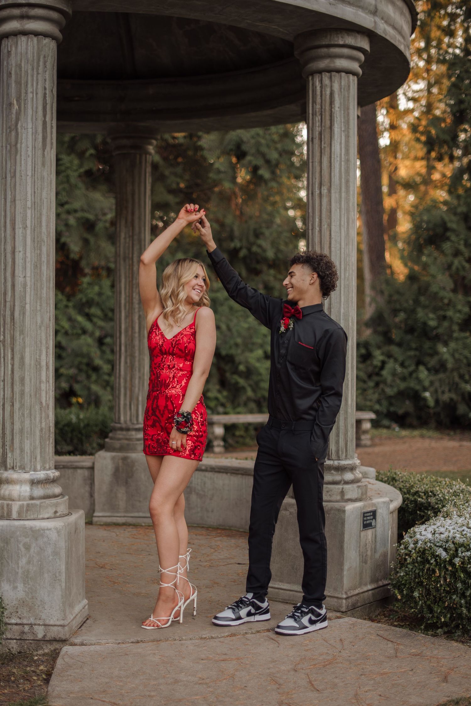 A couple in formal attire poses playfully in a stone gazebo during golden hour.