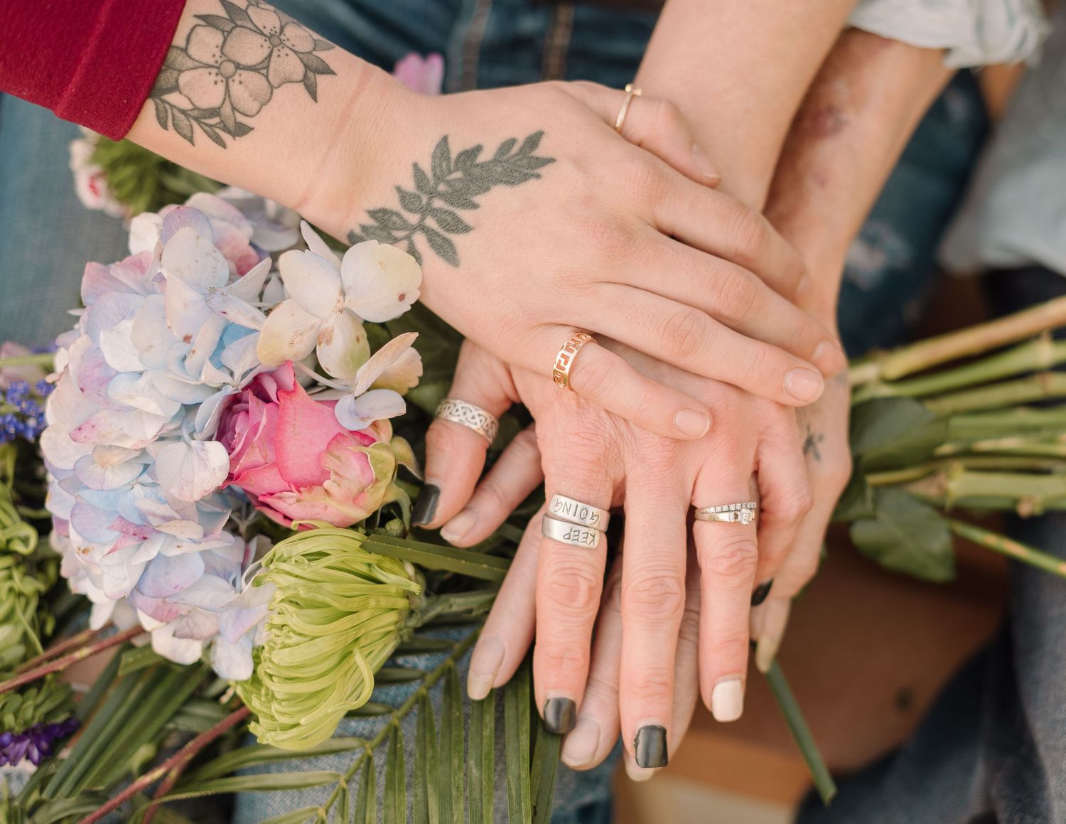 Hands with wedding rings and floral tattoo rest on a bouquet of pink and blue hydrangeas and tropical greenery.
