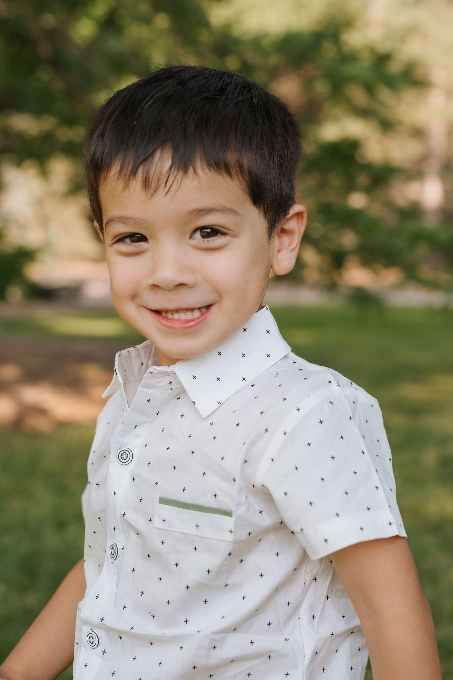 A young child wearing a white polka dot collared shirt sits outdoors on grass with a bright cheerful expression.