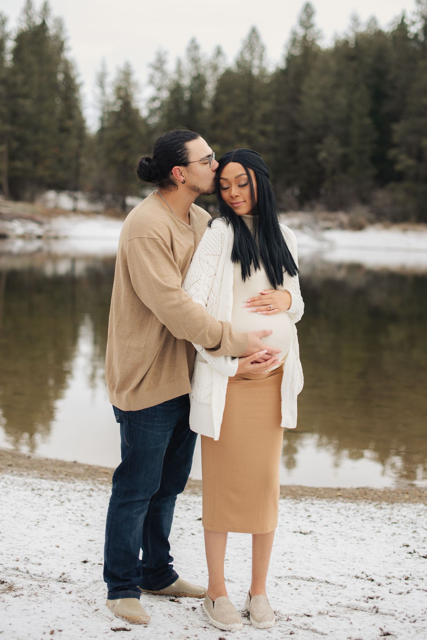Winter engagement photo of couple embracing by snowy lake with evergreen trees.