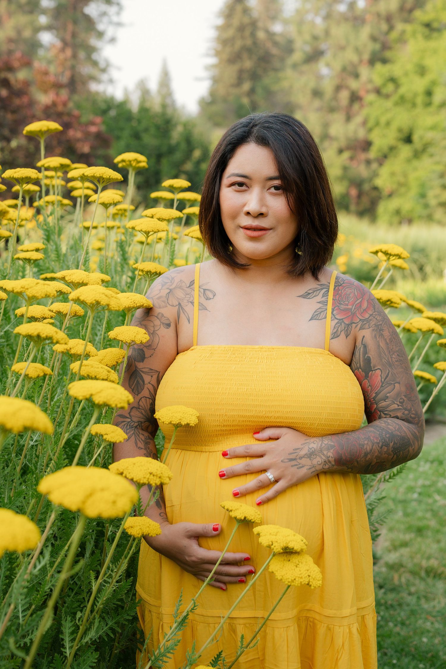 Pregnant woman in yellow dress with tattoos stands among yellow flowers in a lush garden, cradling her baby bump.