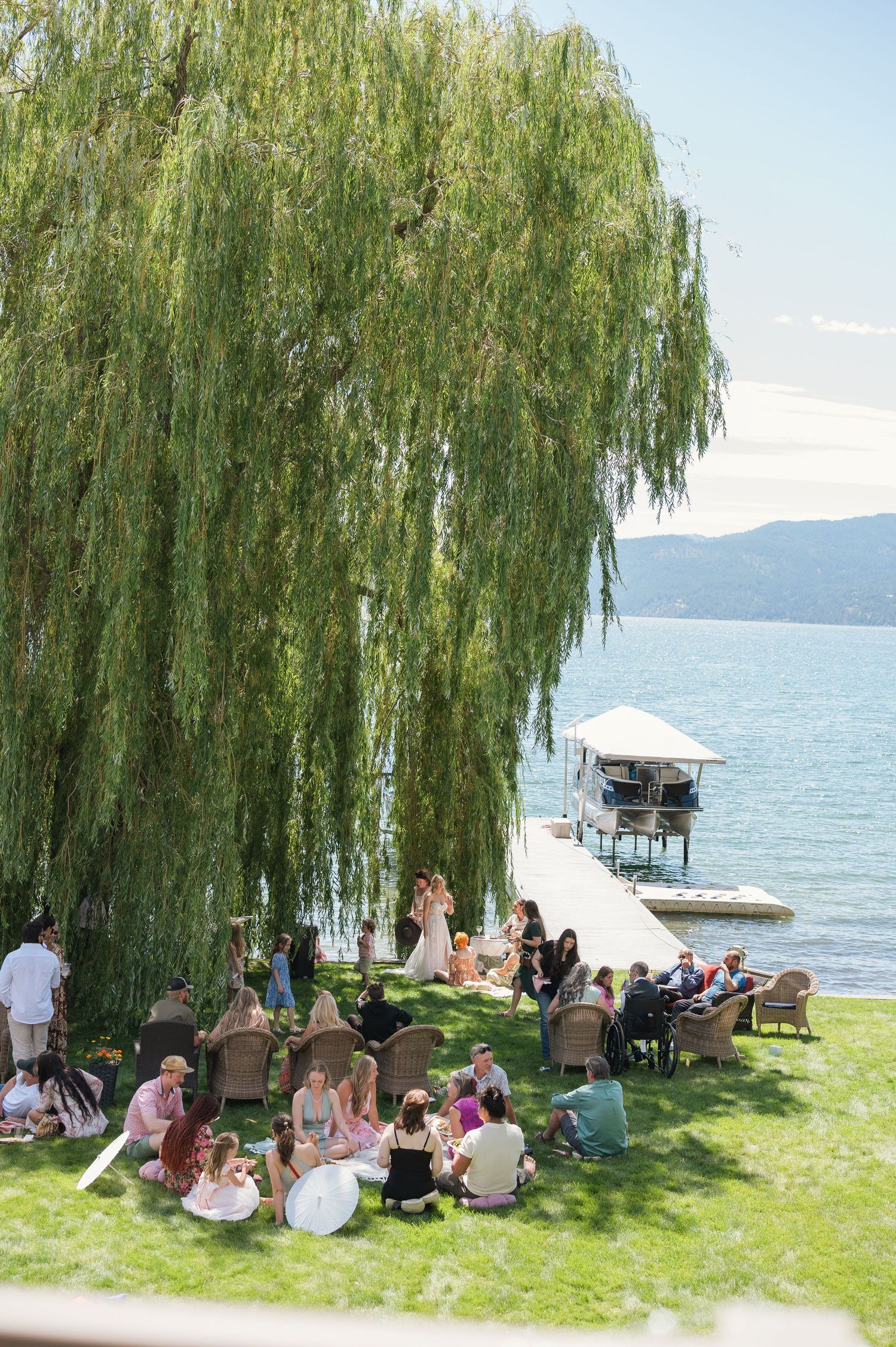 Guests gather under a weeping willow tree by a lakeside dock on a sunny day for an outdoor celebration.