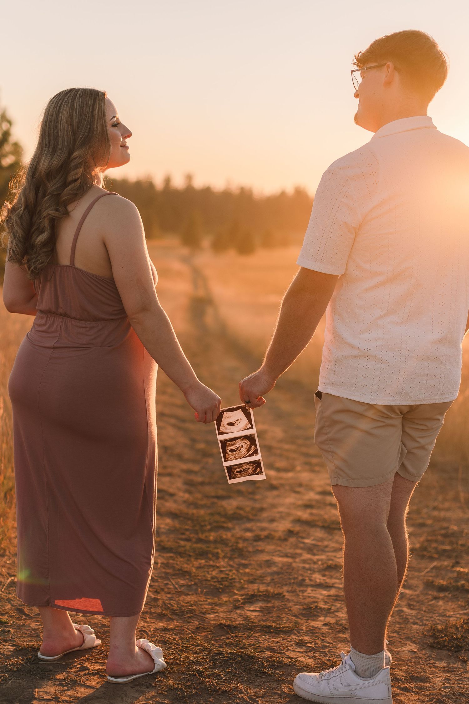 A couple holding ultrasound photos stands together at sunset in a golden field wearing casual attire.