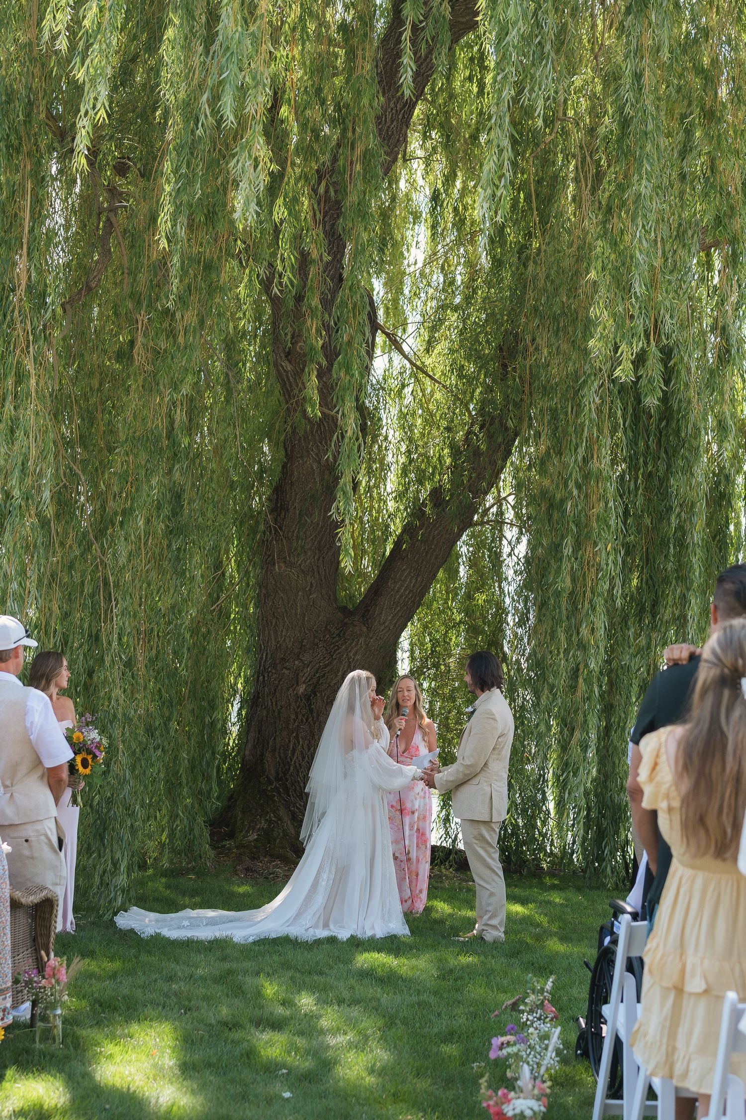 Outdoor wedding ceremony under a weeping willow tree, with bride in white gown and veil exchanging vows.