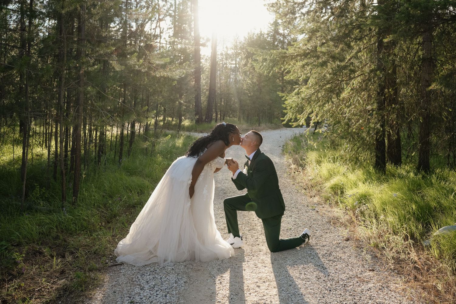 Bride and groom sharing a romantic kiss on a forest path, groom kneeling in a dark green suit, sunlight filtering through tall pine trees.