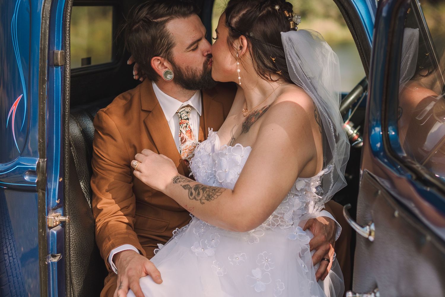 A couple shares a romantic kiss while seated in a vintage blue car during their wedding celebration.