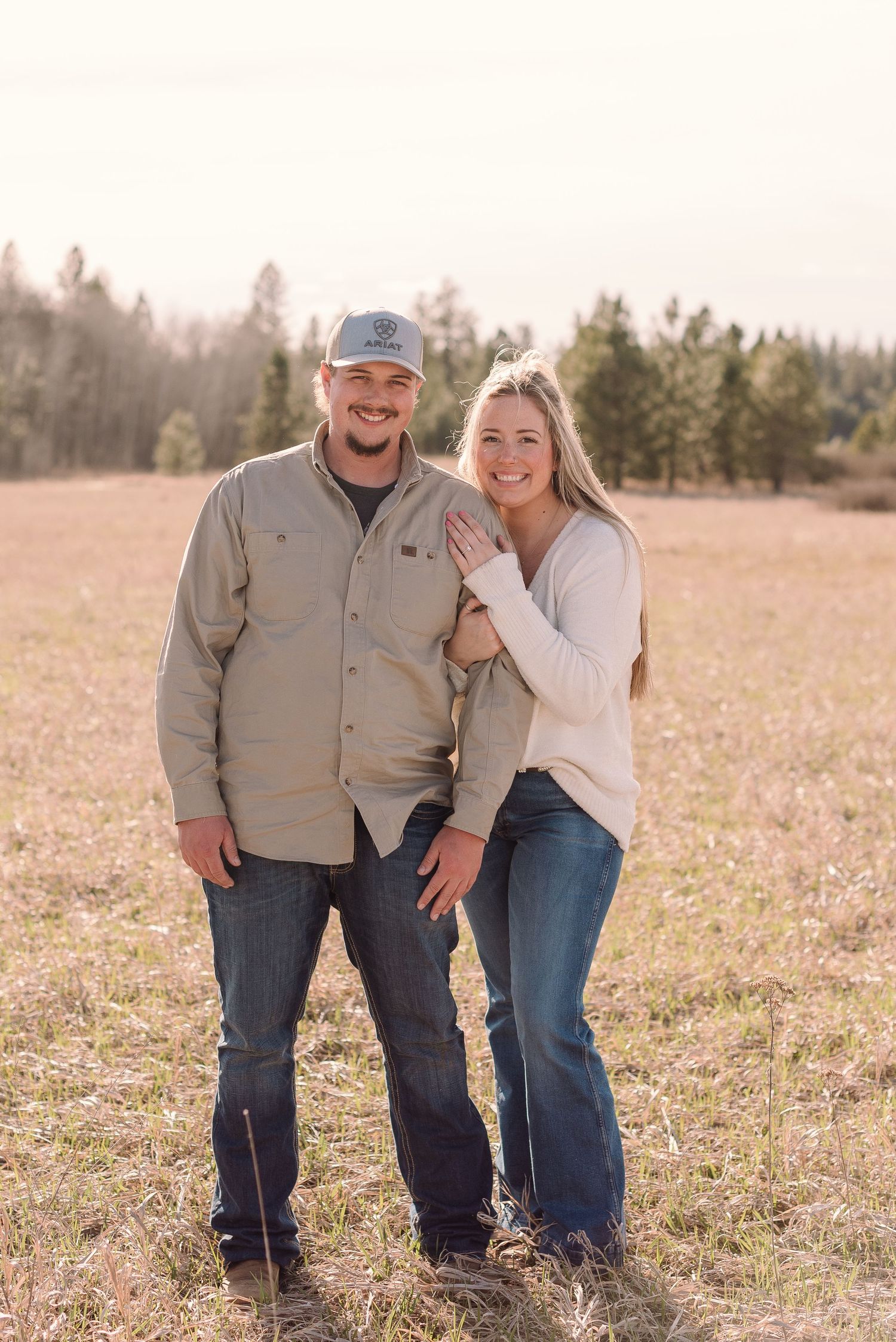 A couple poses together in a sunny field with evergreen trees in the background during an outdoor photo session.