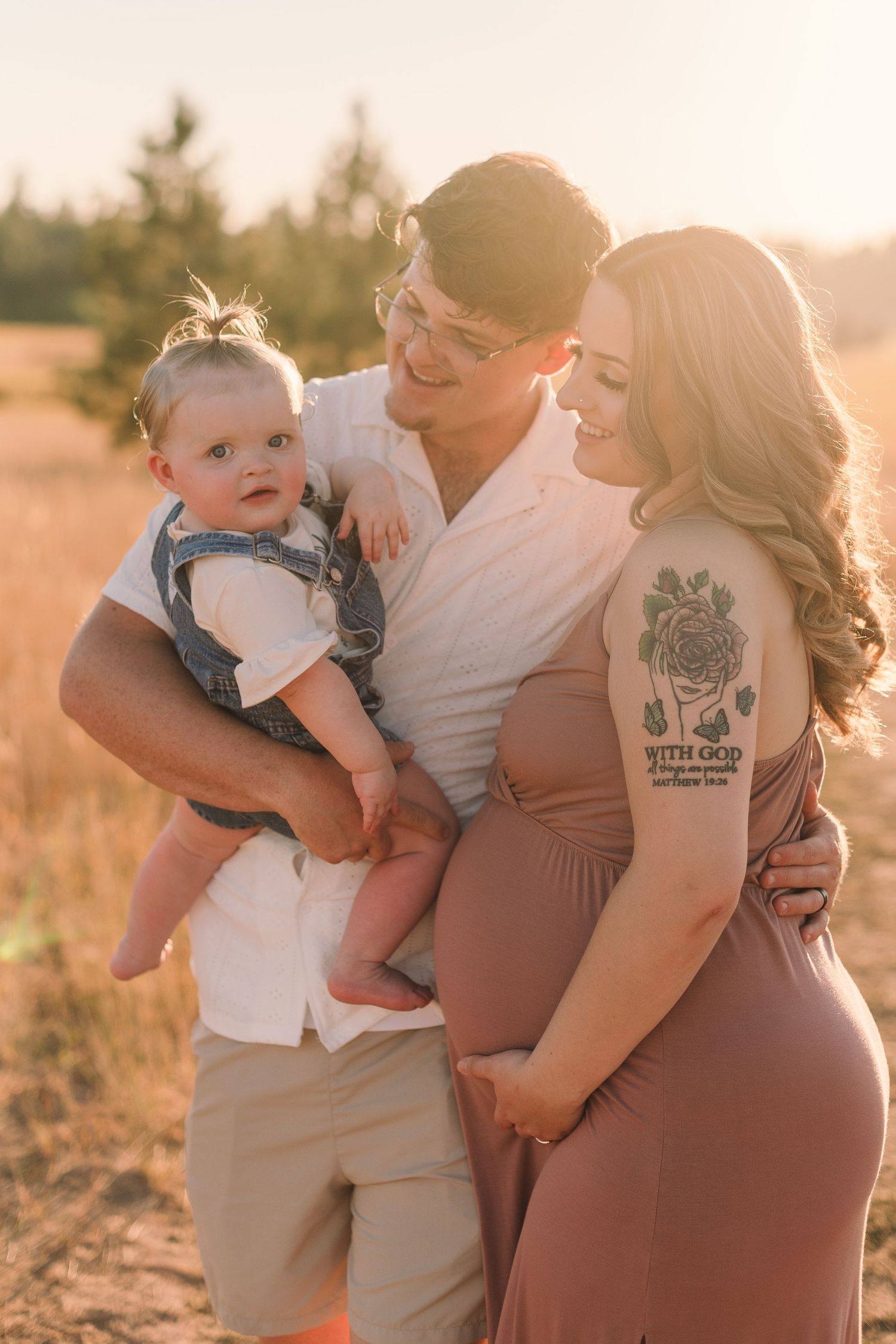 A family poses together in a golden sunset field, with a baby and a pregnant woman in a mauve dress.