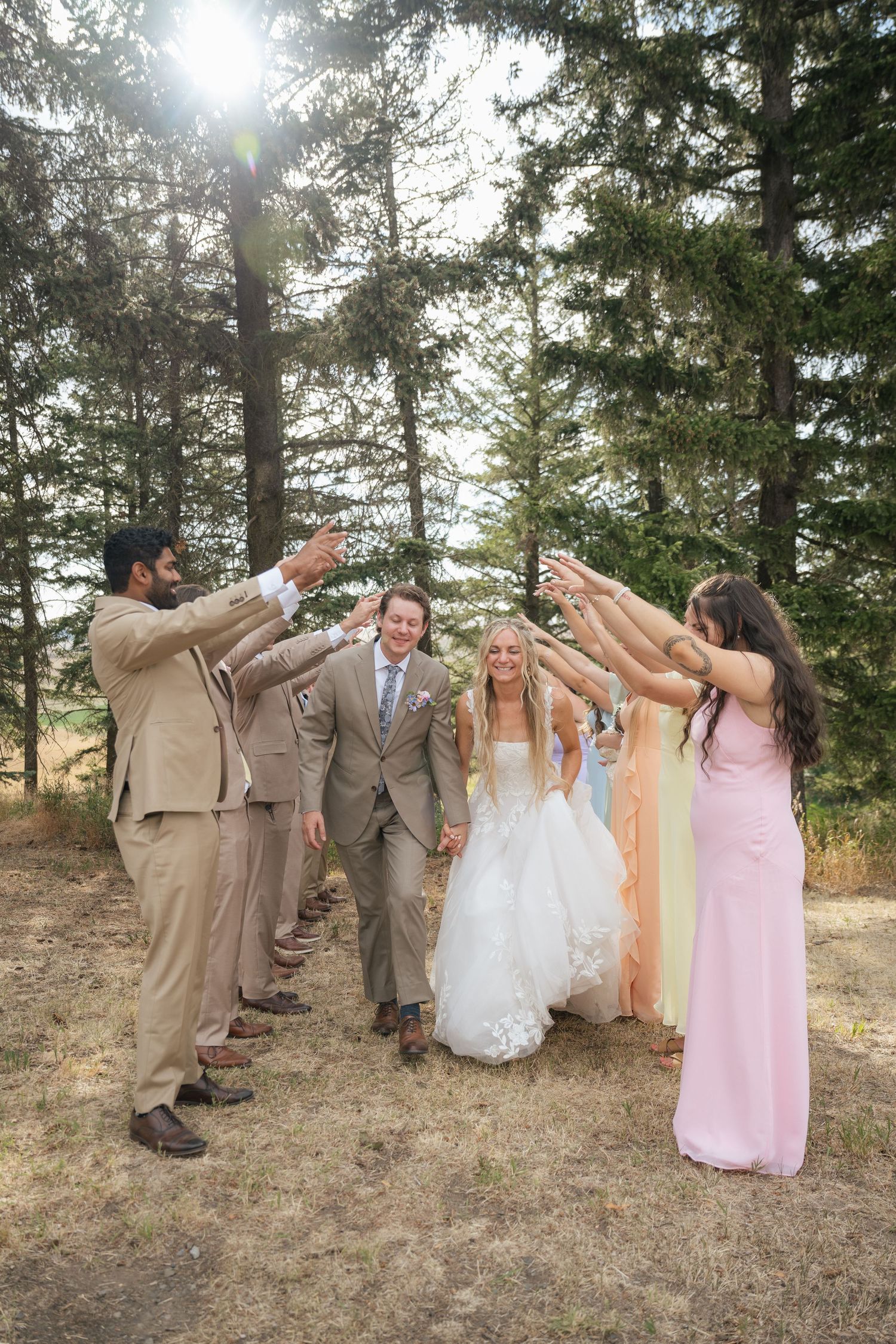 Bride and groom walk through arch of raised hands formed by wedding party in tan suits and pastel dresses outdoors.
