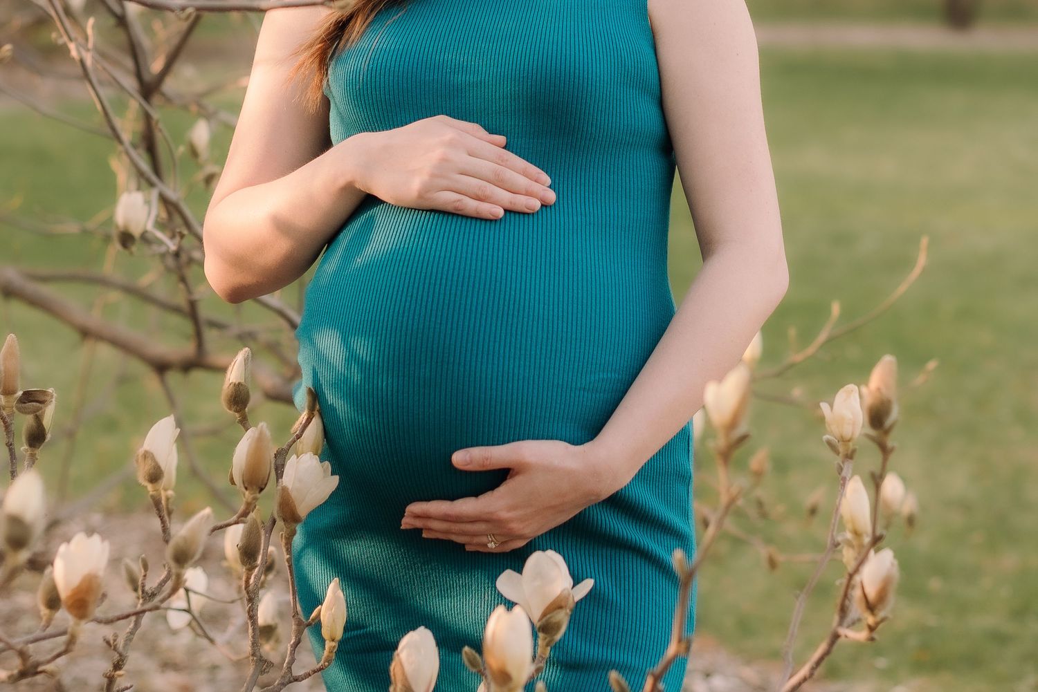 A pregnant woman in a teal dress cradles her baby bump while standing in a field with autumn foliage.