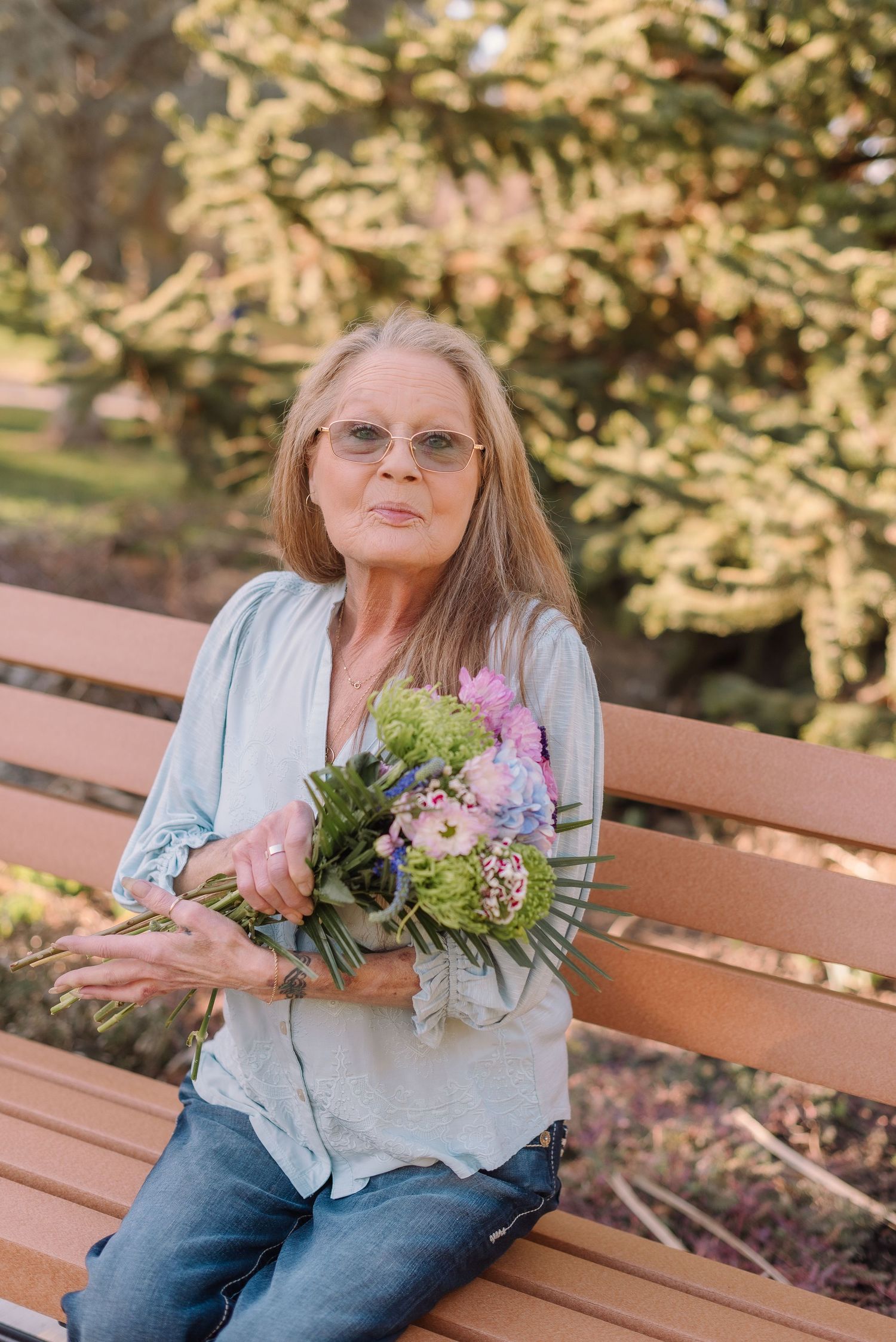 A person sits on a wooden bench outdoors holding a colorful bouquet of pink and white flowers in casual attire.