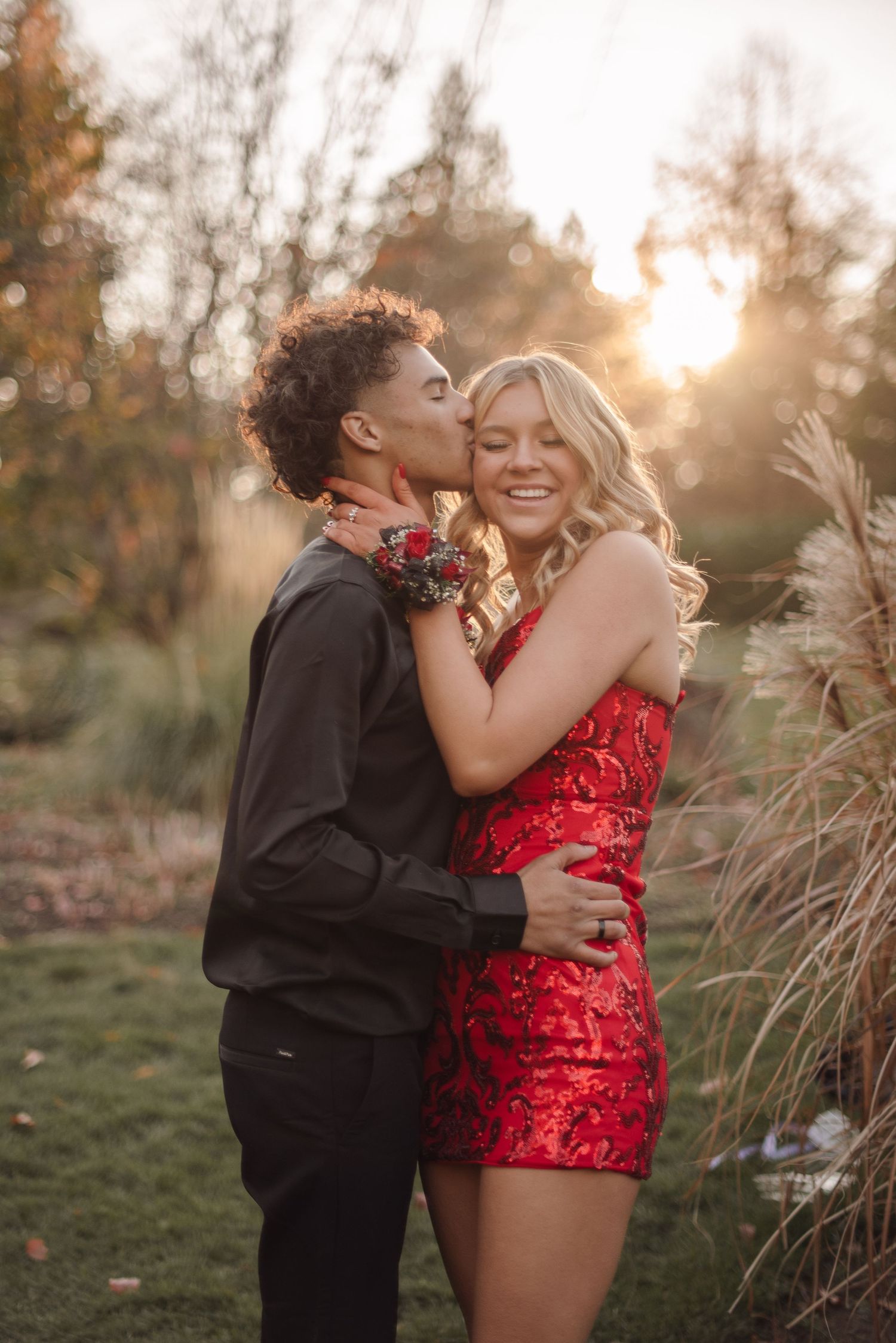 A couple embraces in a sunlit field with tall grass during golden hour while wearing red and black outfits.