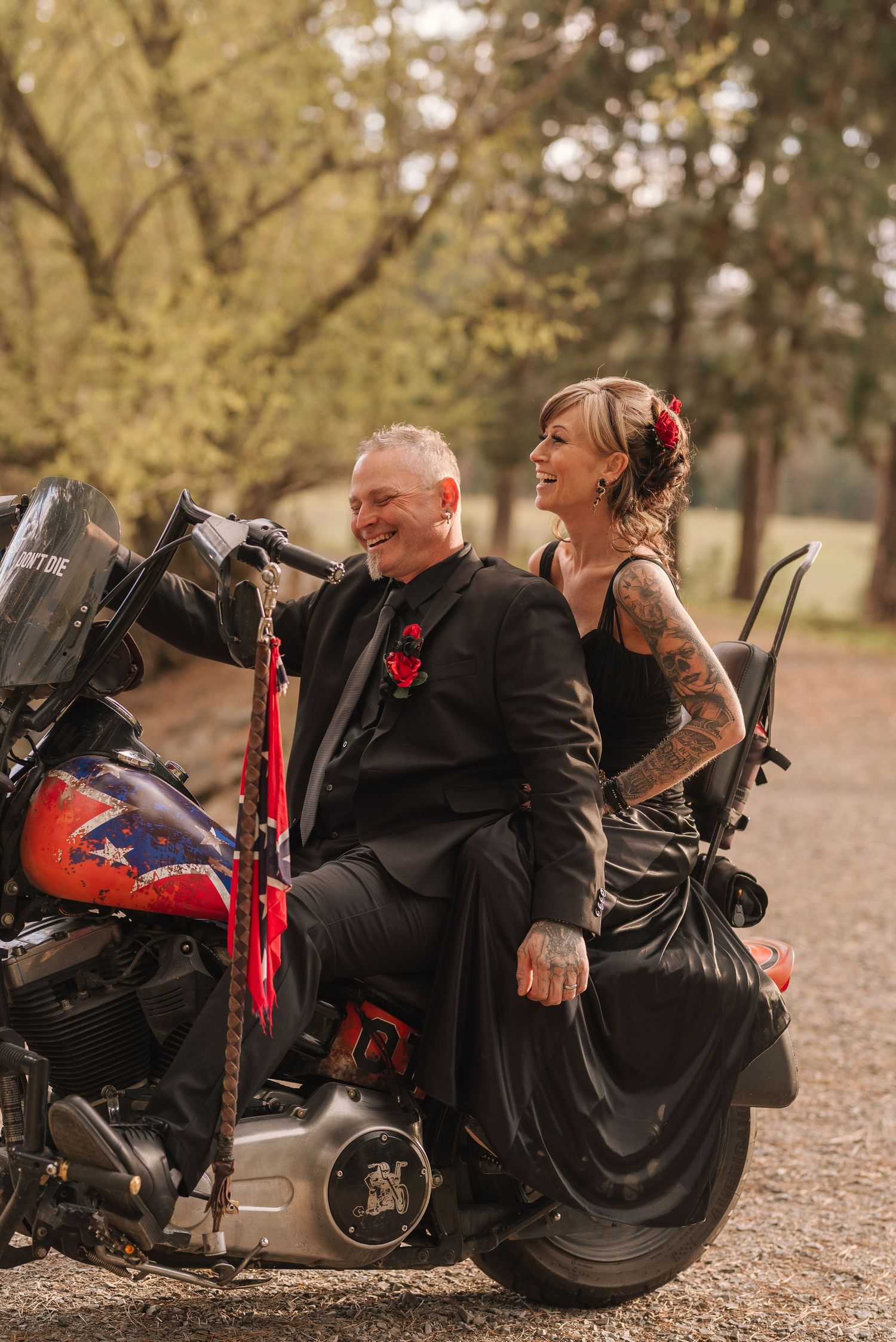 A couple in black attire share a romantic moment while sitting on a motorcycle on a leaf-covered ground.