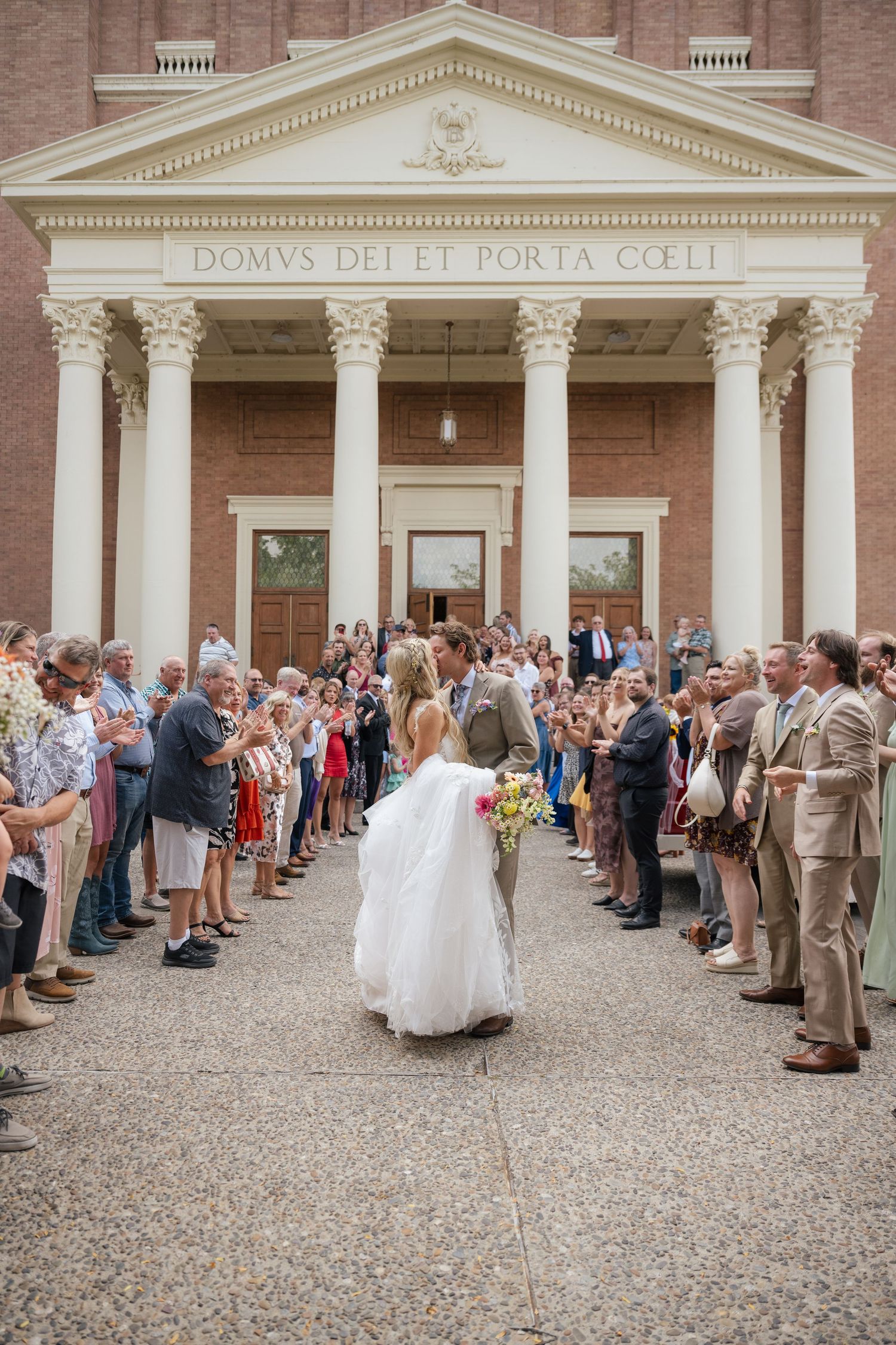 Bride and groom kiss outside a brick church with Latin inscription, surrounded by cheering wedding guests.