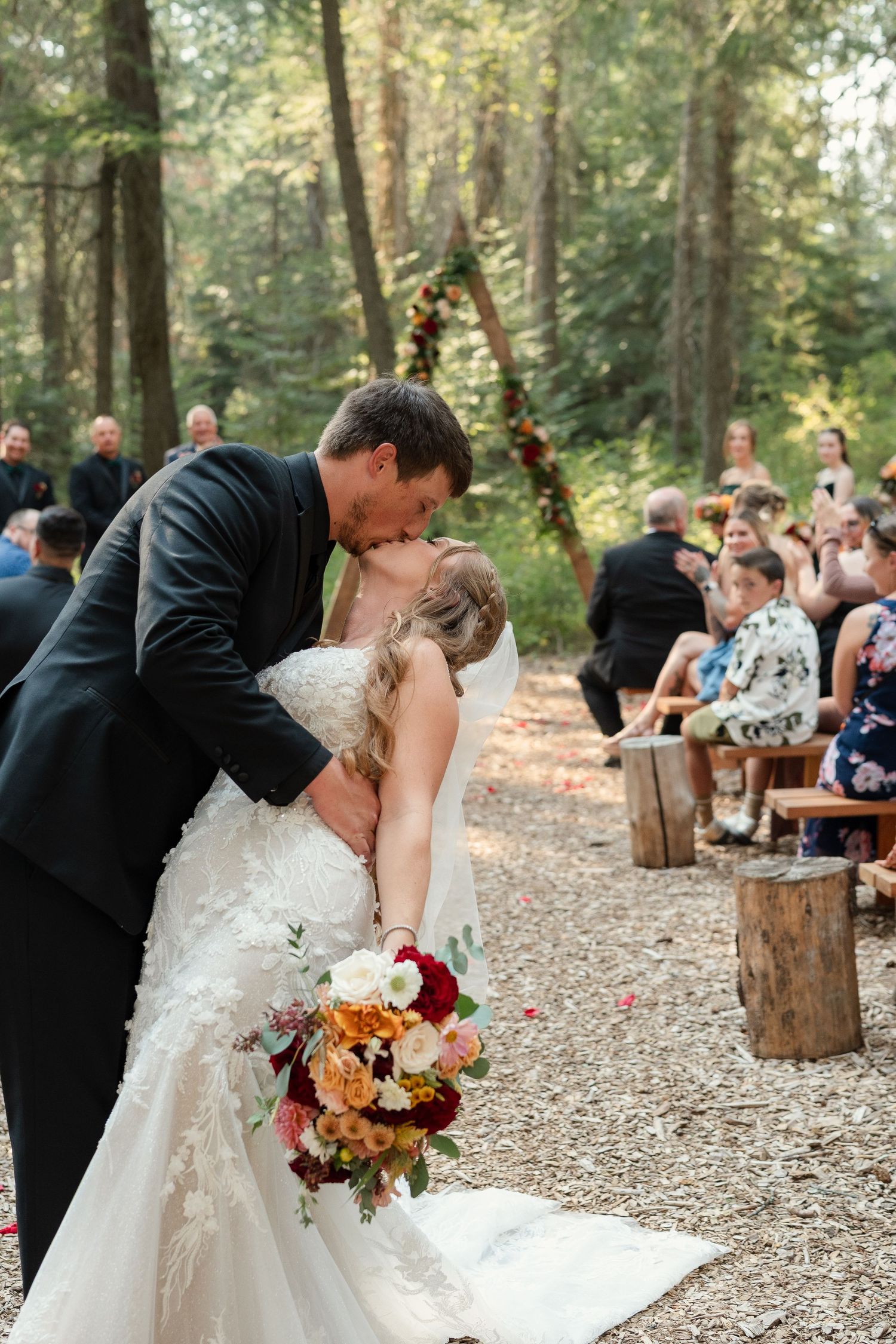 Bride and groom share first kiss at outdoor forest wedding ceremony with floral arch and seated guests.
